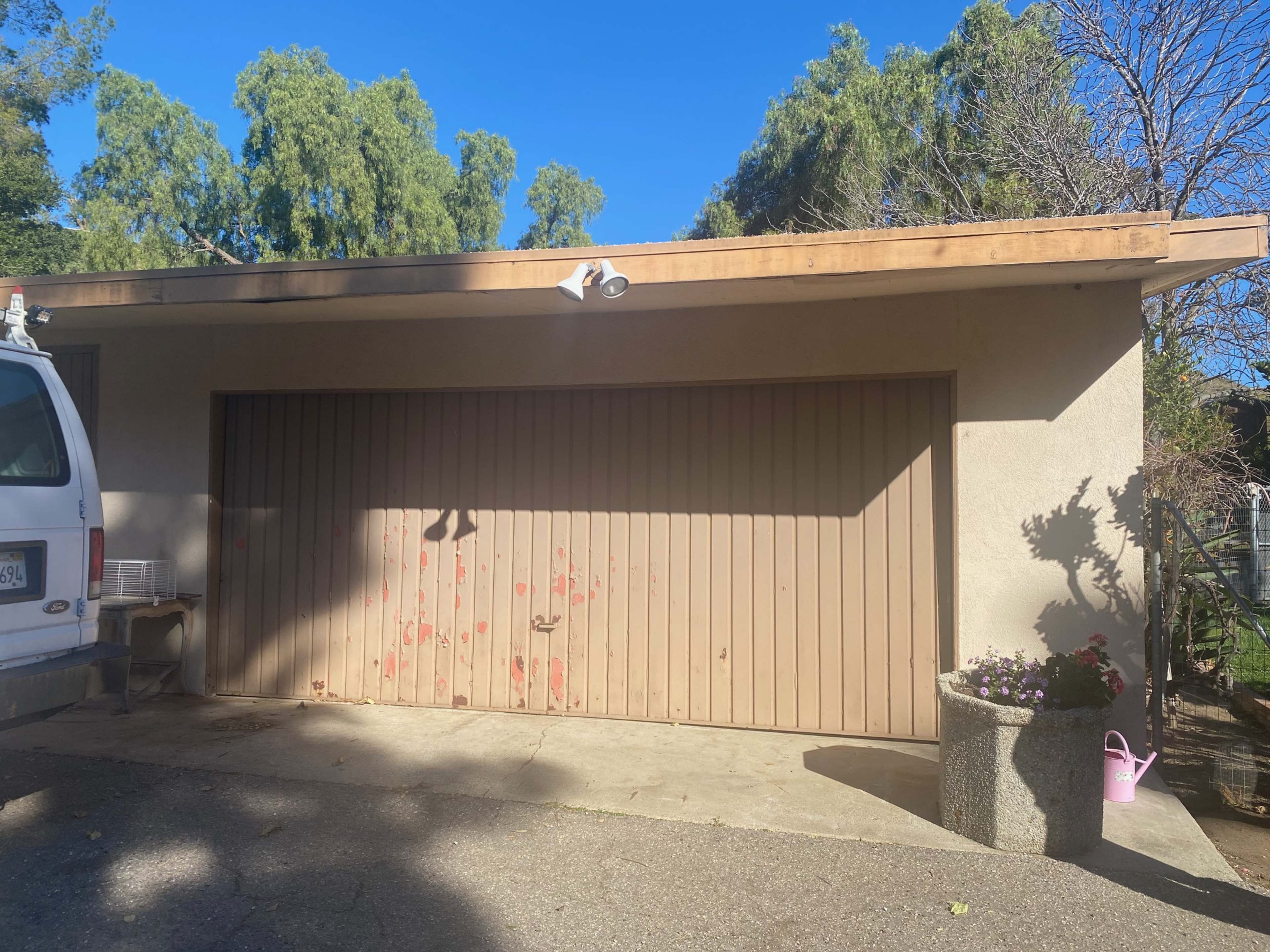A garage with a closed door, featuring weathered paint and a flower planter nearby, is set against a clear blue sky.