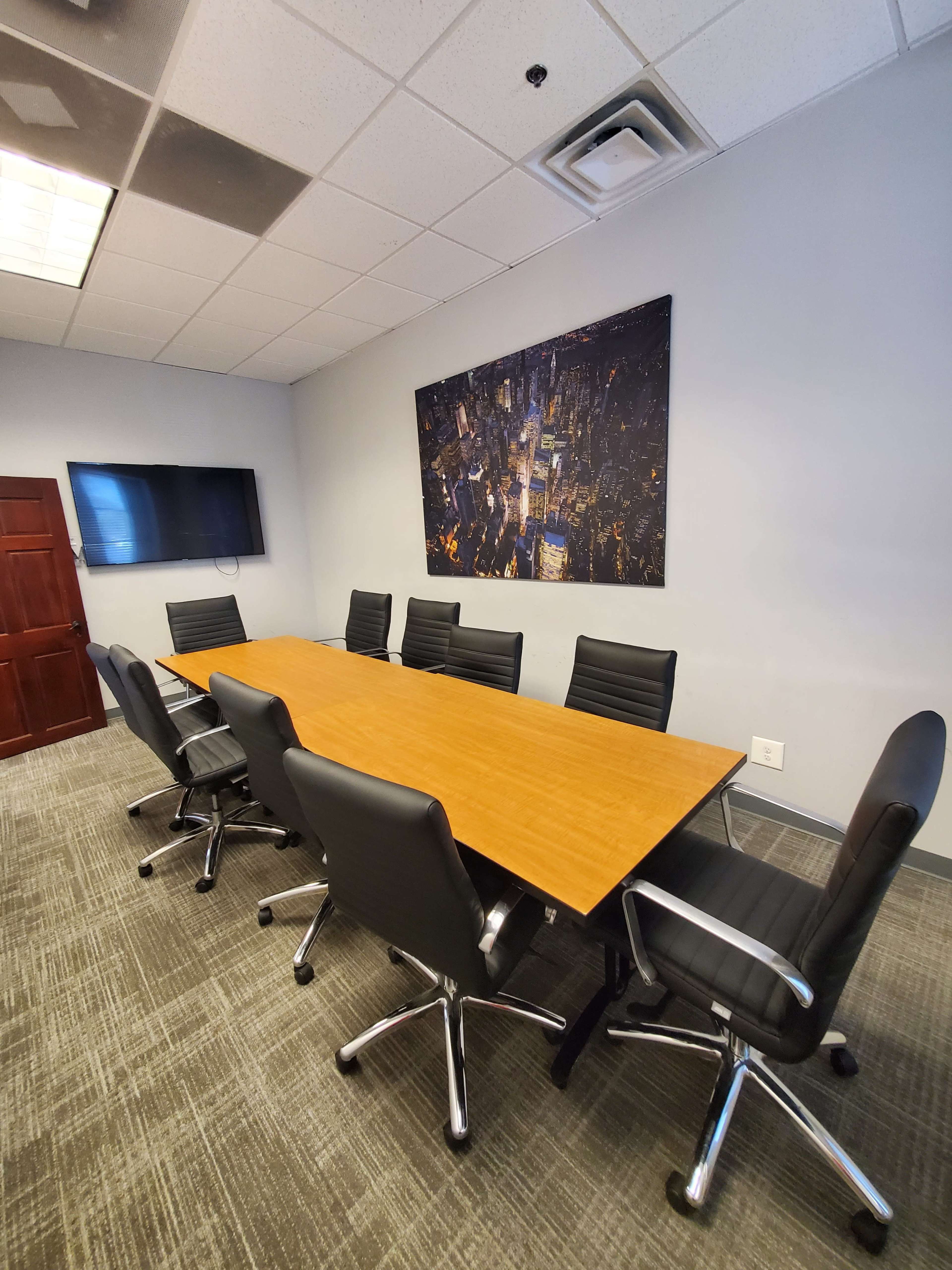 A modern conference room with a long wooden table surrounded by black swivel chairs, a large wall-mounted screen, and a cityscape picture on the wall.