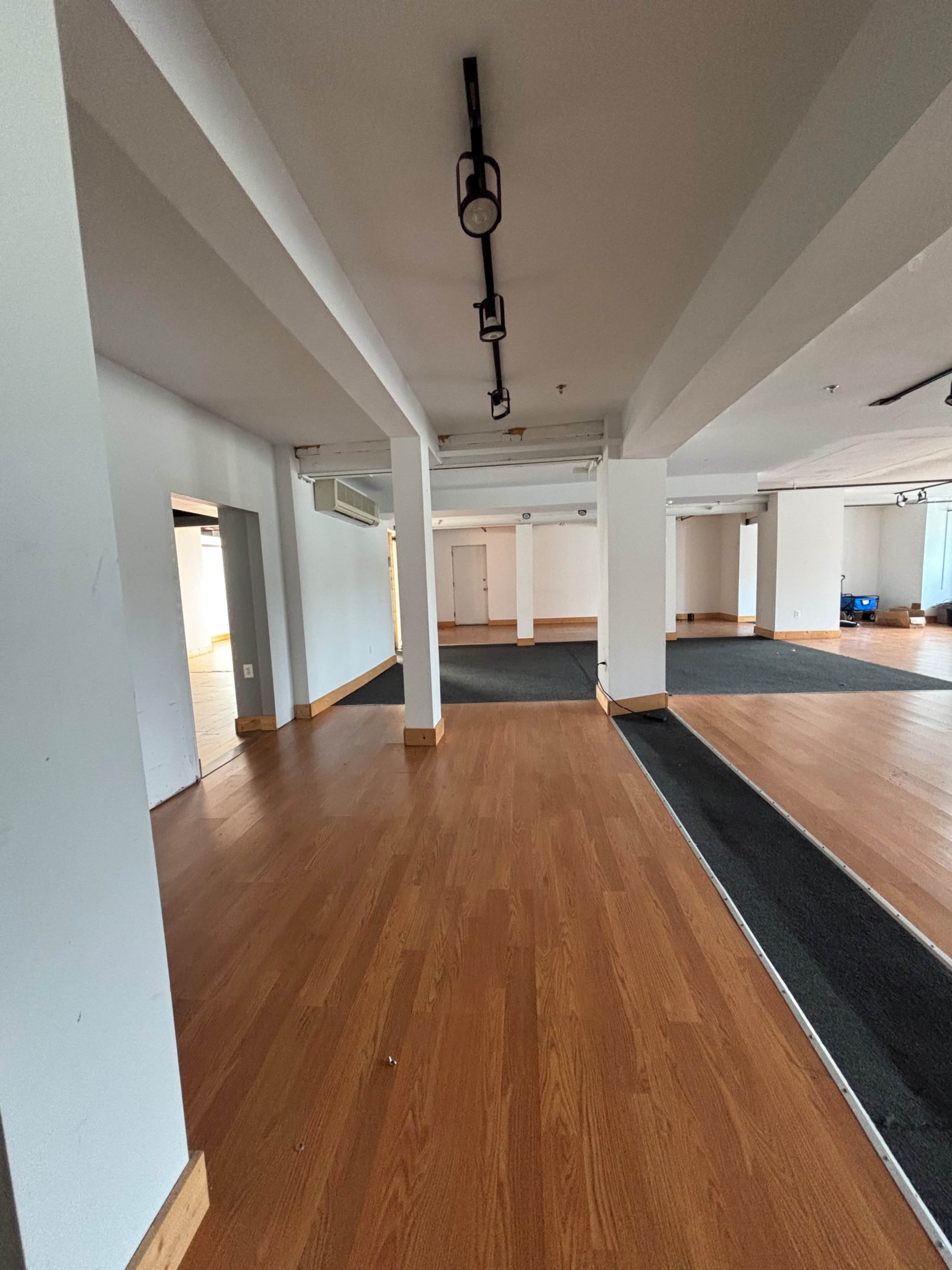 The image shows an empty interior space with wooden flooring, white walls, and several columns.
