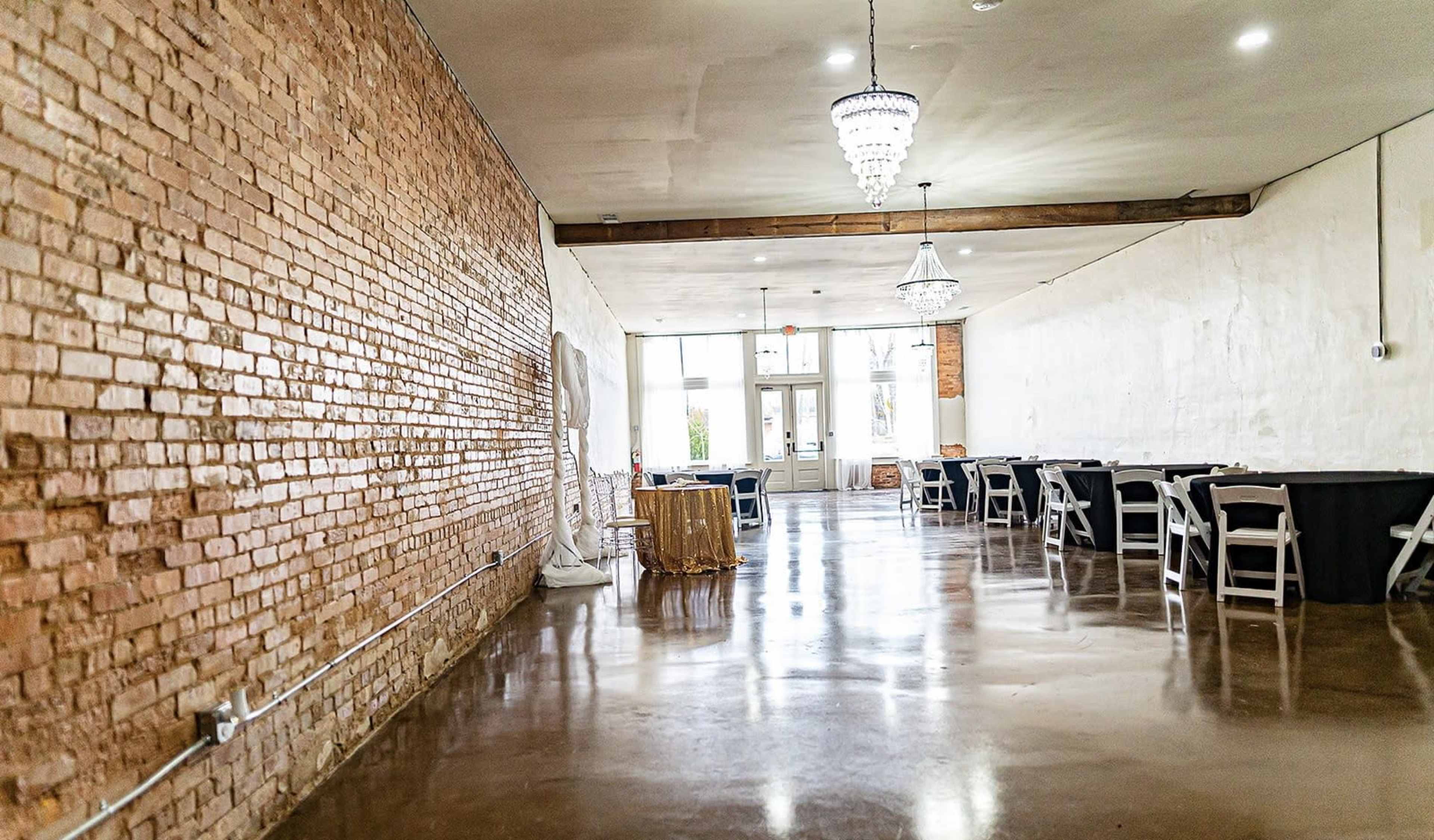 The image shows a spacious event venue with exposed brick walls, polished concrete floors, and several tables set for dining along one side.