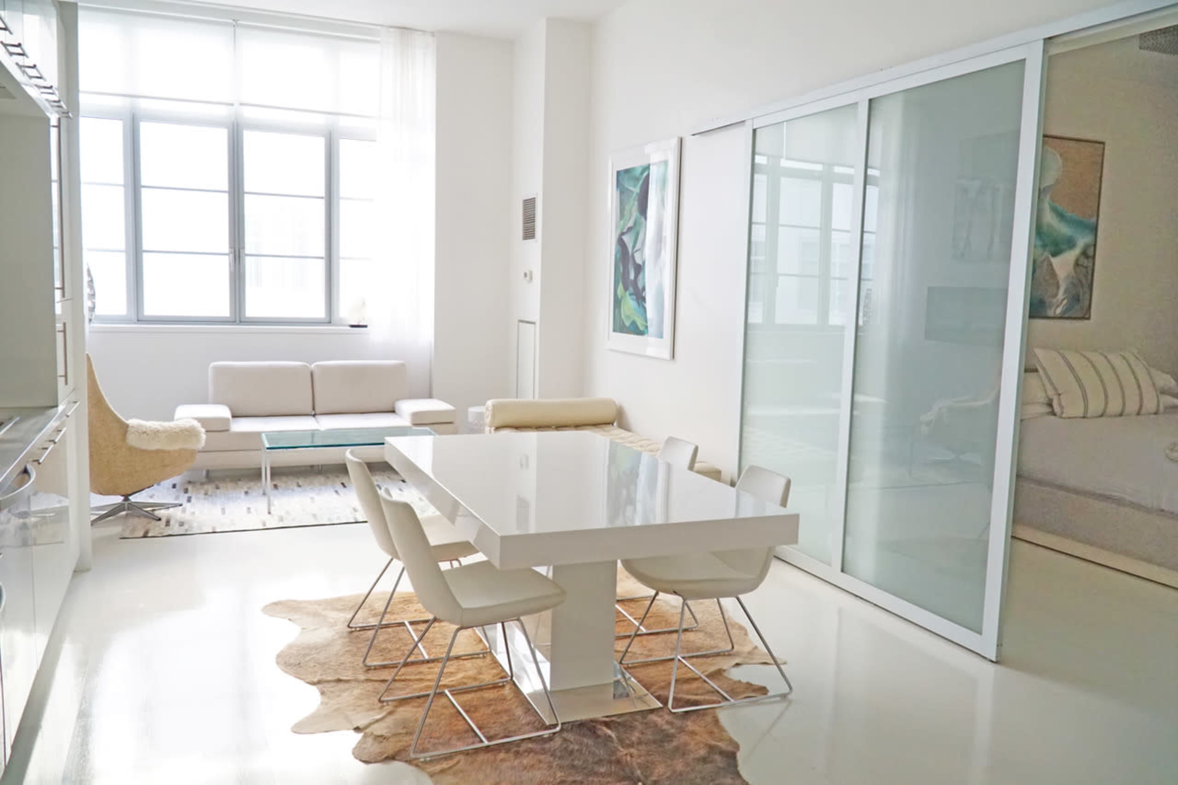 A modern dining area features a white table surrounded by four chairs, with a sofa and large windows in the background.