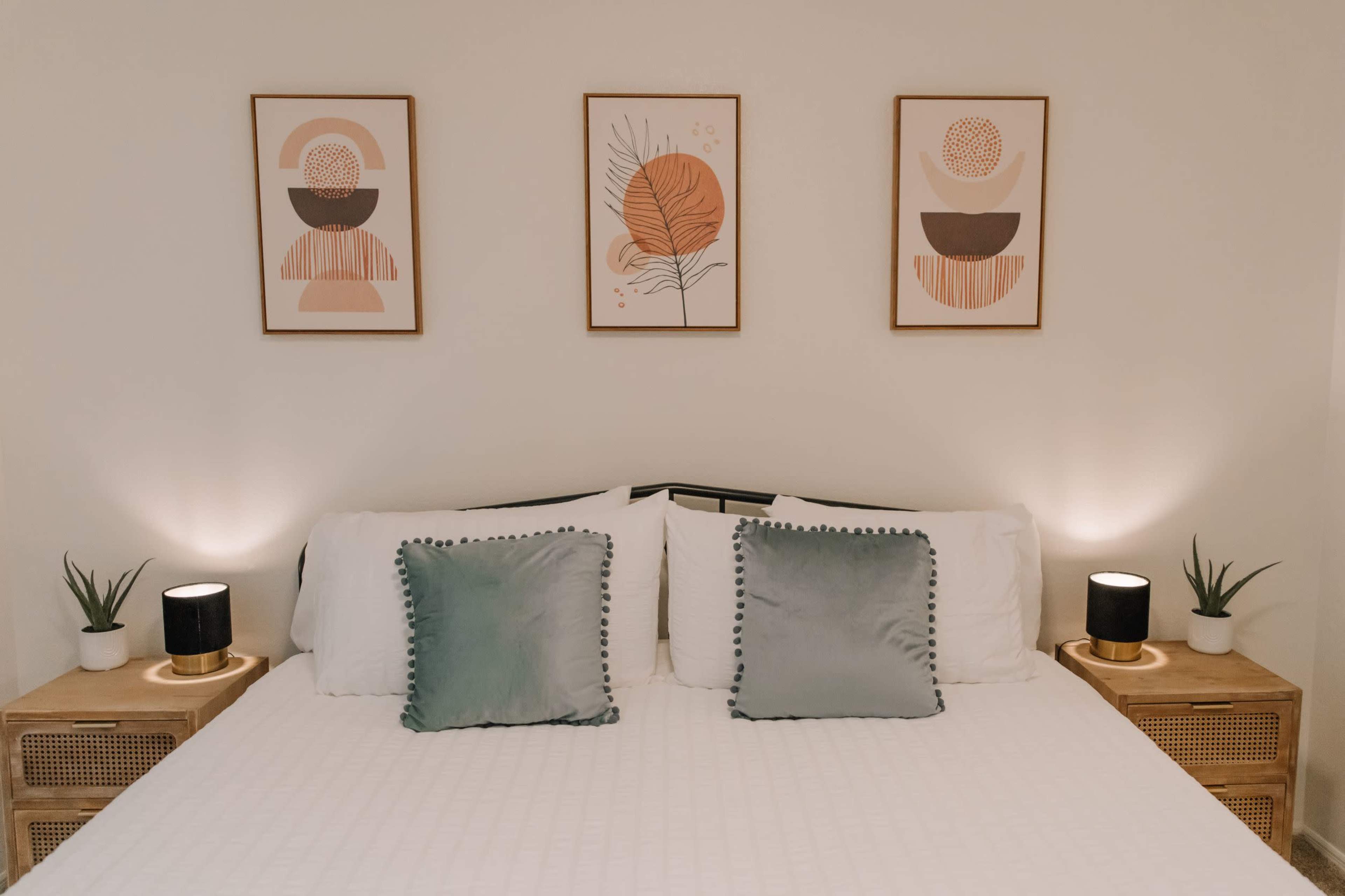 A neatly arranged bedroom with a bed featuring white linens, two decorative pillows, and framed art above the headboard, along with nightstands and small plants on either side.