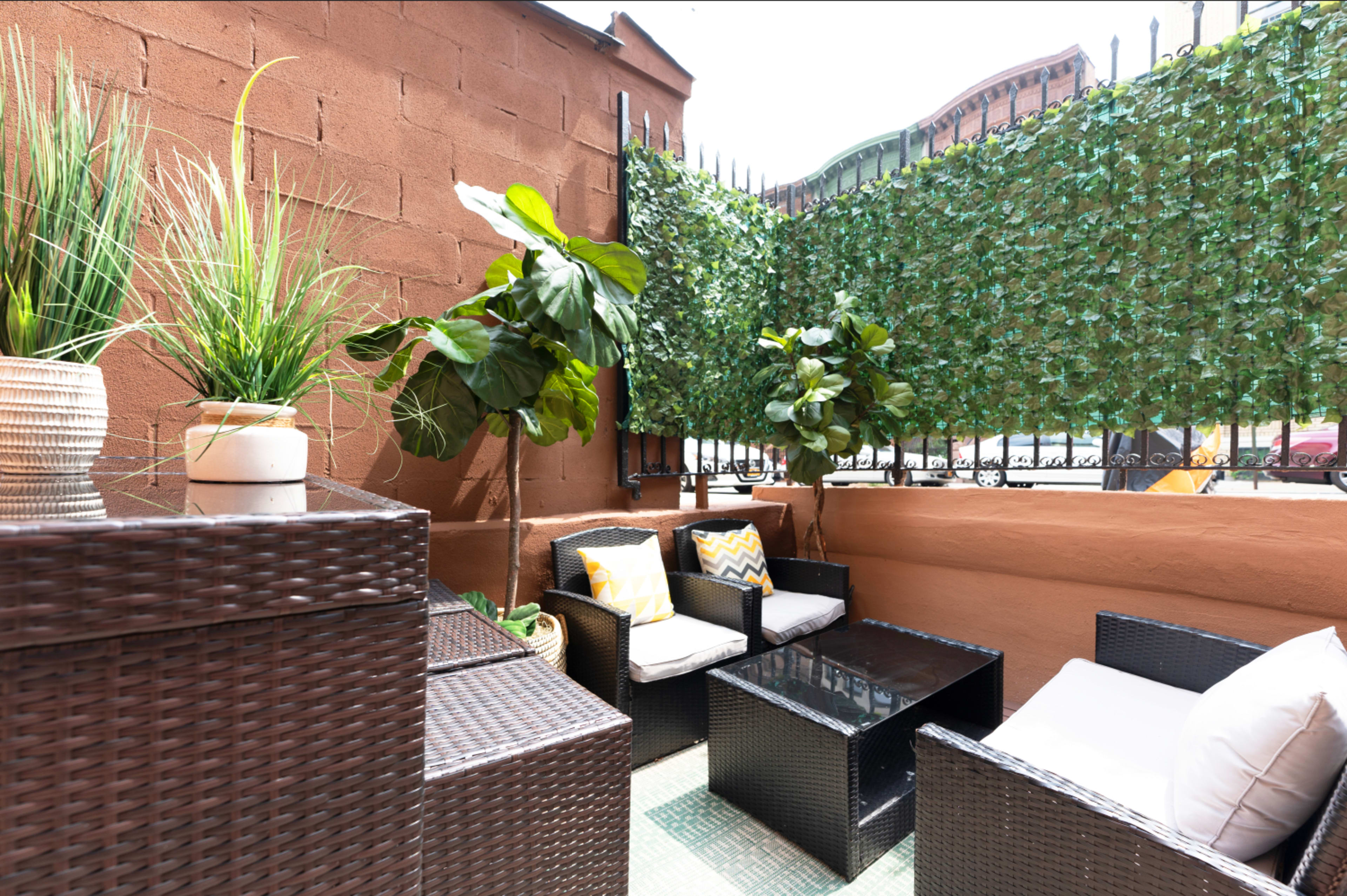 A small outdoor patio area with wicker furniture, potted plants, and a green leafy privacy screen.