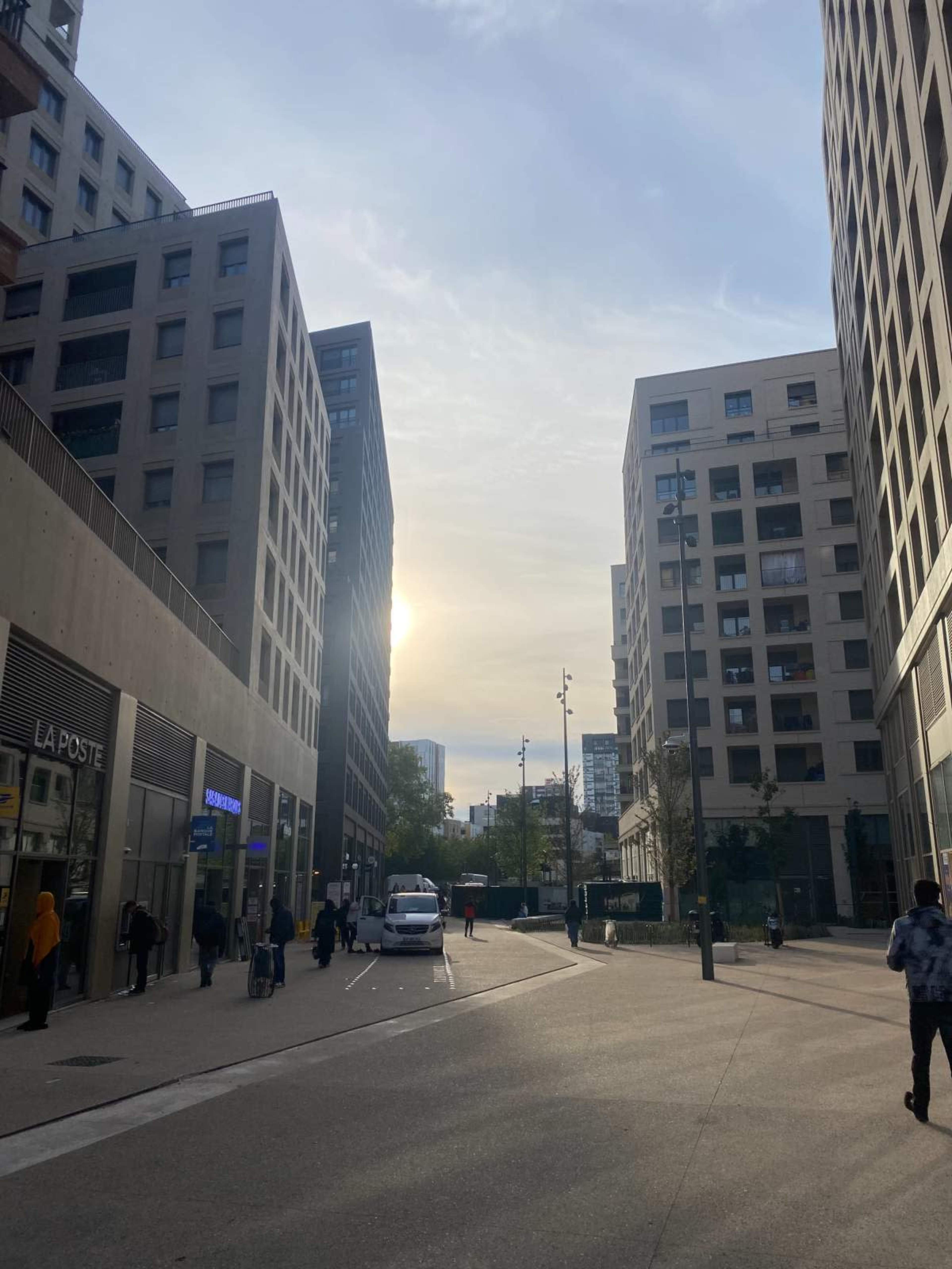 The image shows a modern urban street lined with rectangular buildings, with a vehicle parked near a shop and a hint of sunlight peeking through the gap between the structures.