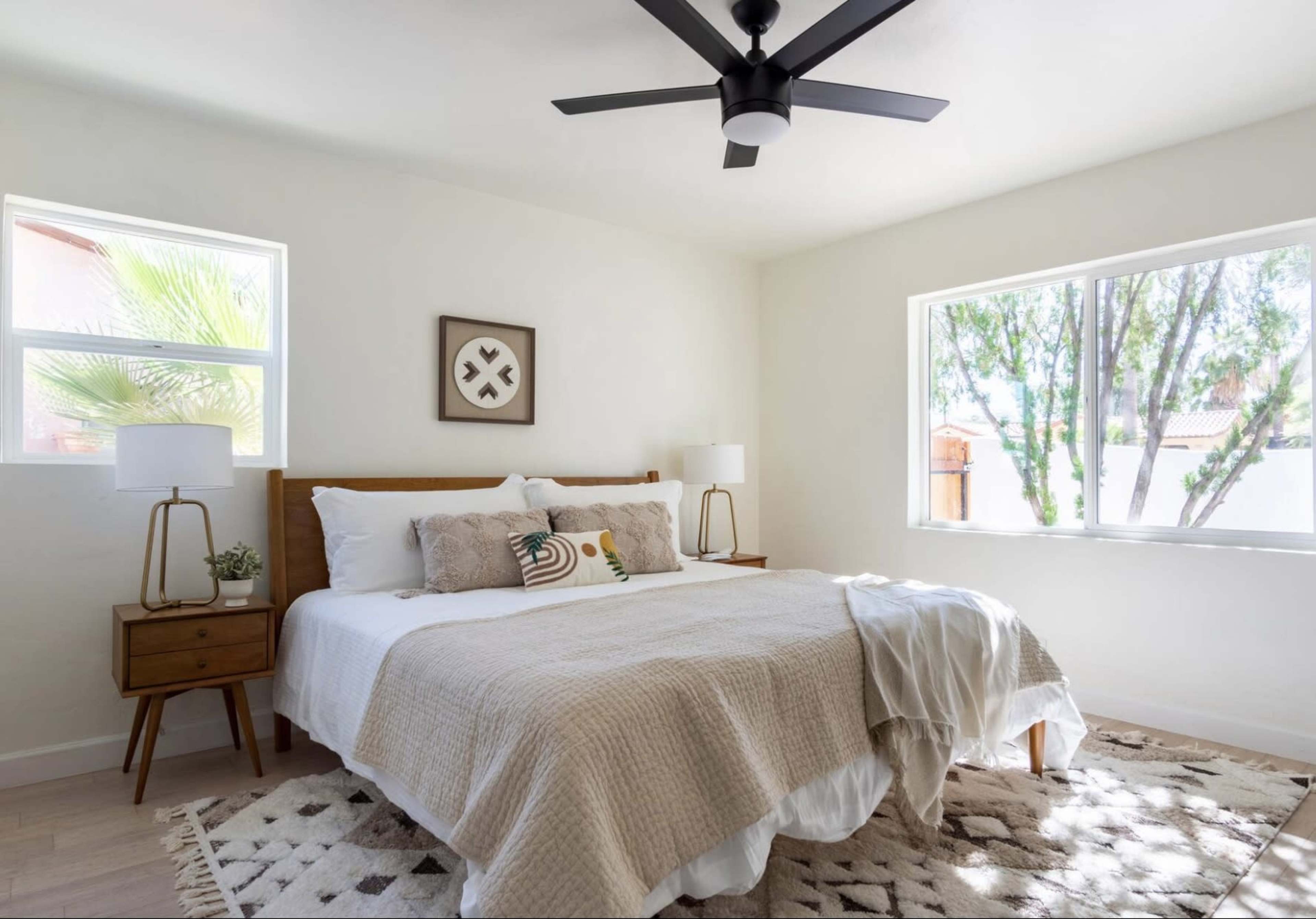 A well-lit bedroom features a large bed with neutral bedding, wooden side tables with lamps, and windows that overlook greenery outside.