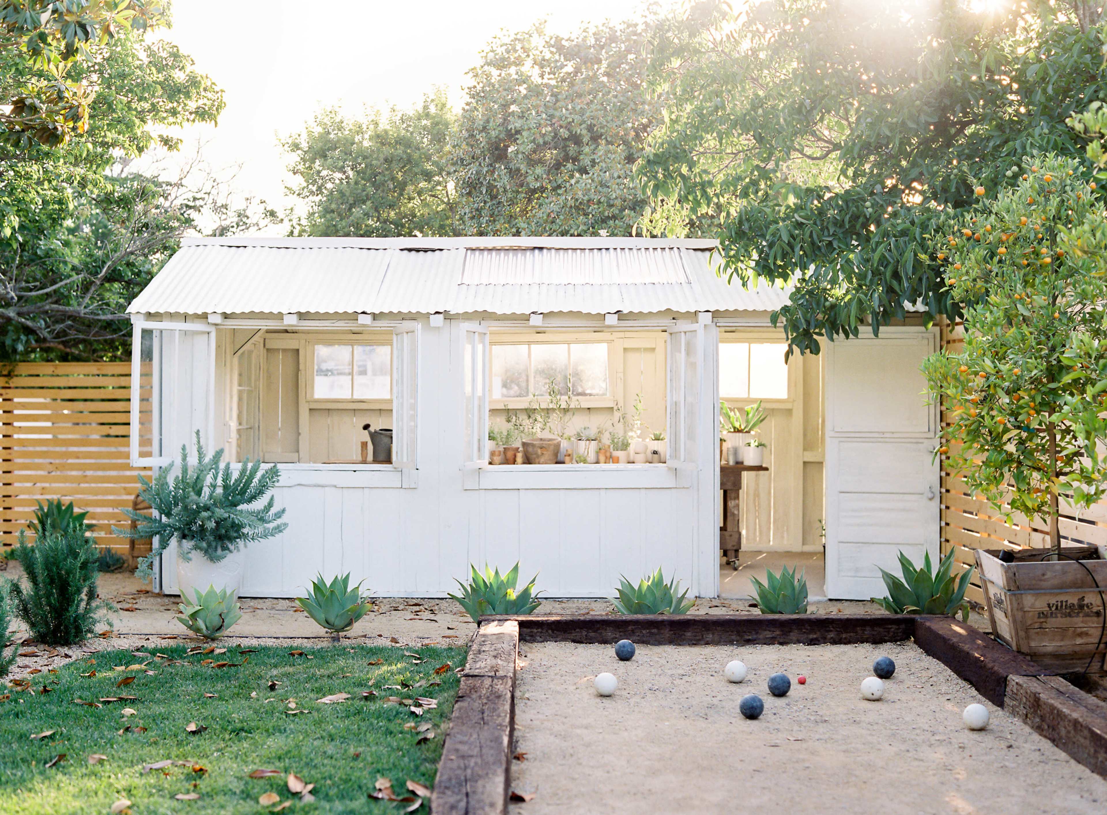 A white shed with large windows stands in a garden, next to a sand court with several balls arranged.