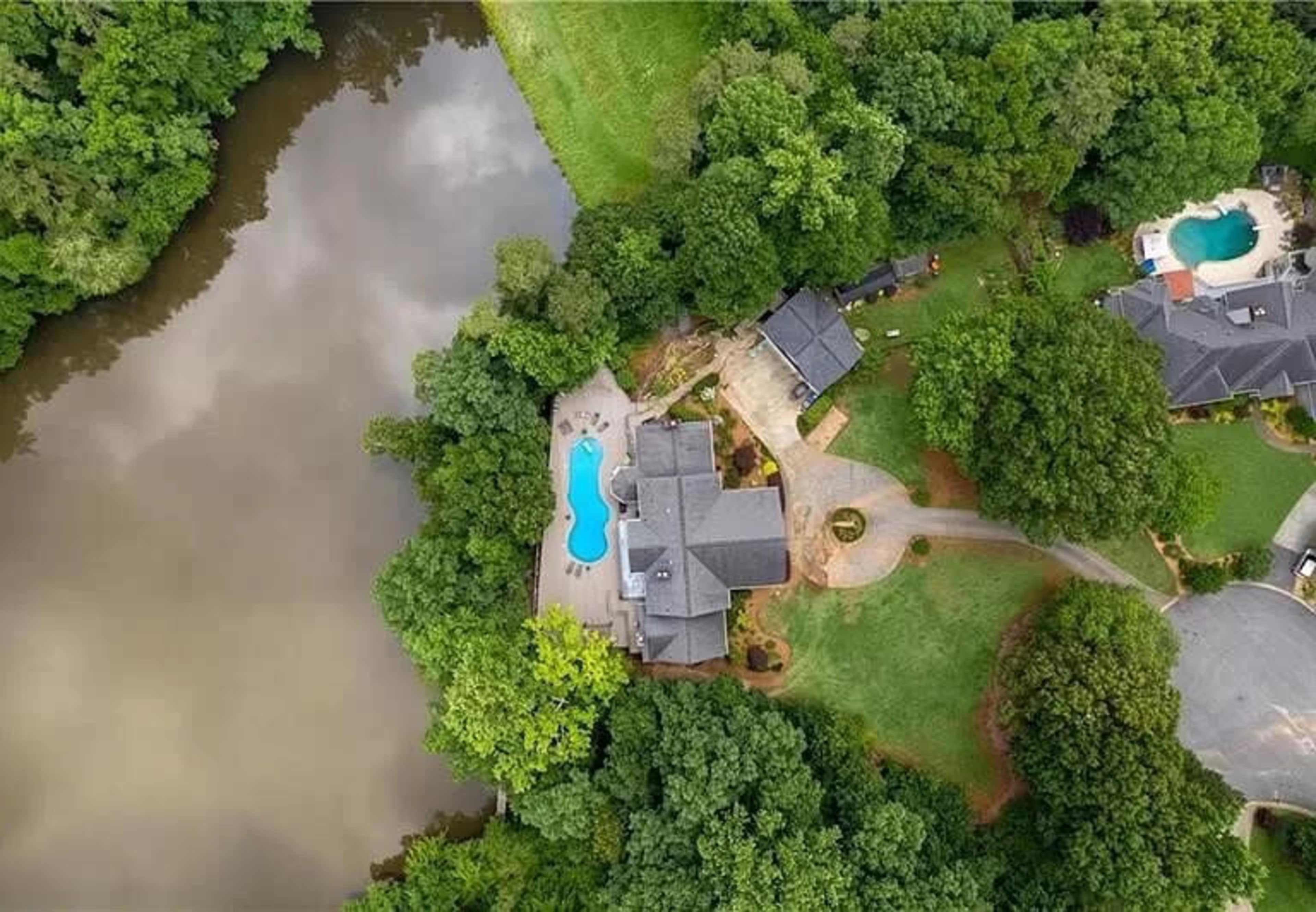 An aerial view shows a large house with a swimming pool surrounded by trees and a river nearby.