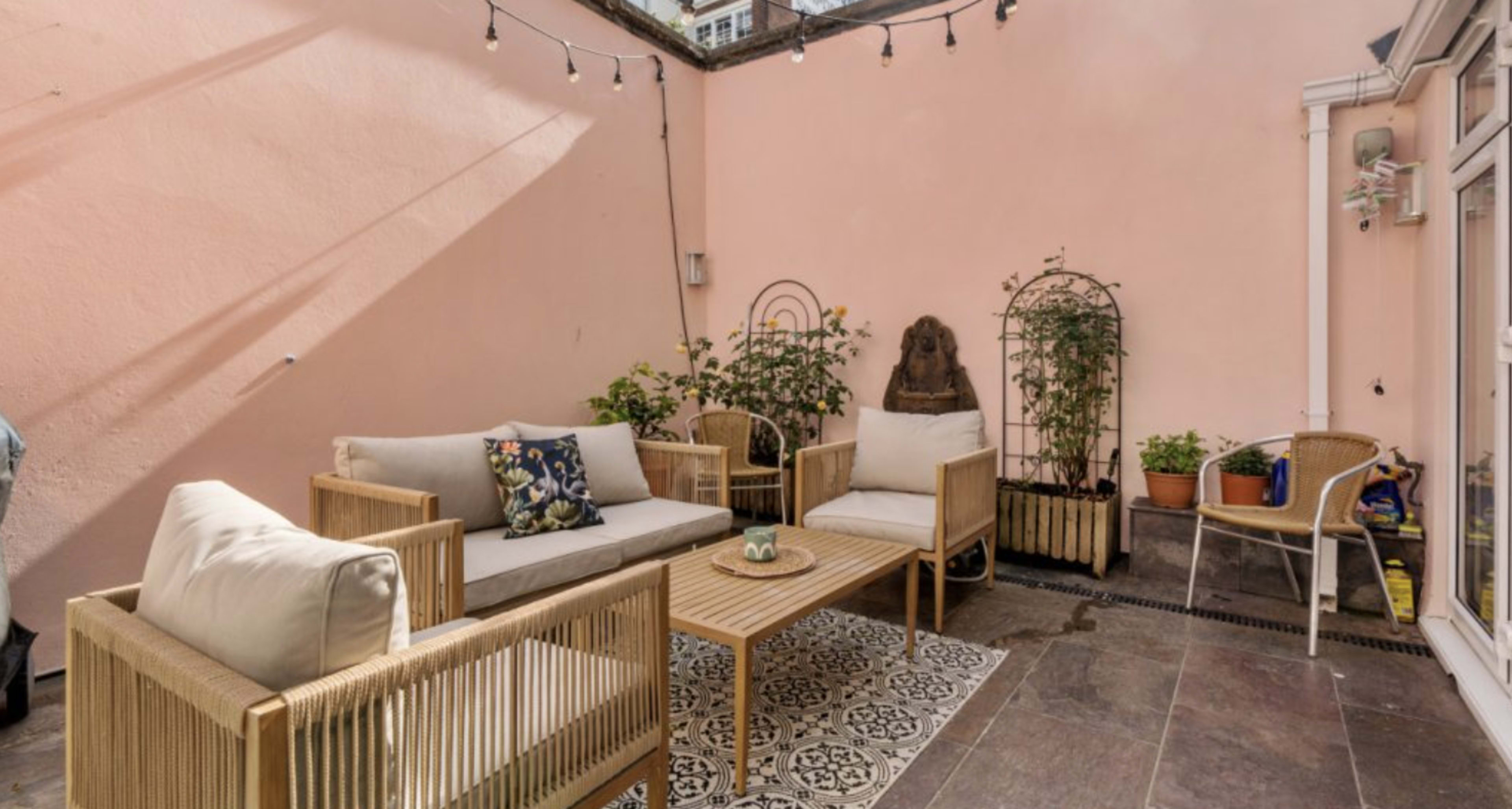 A small outdoor patio features a seating area with wicker furniture, a central wooden coffee table, and potted plants against a light pink wall.