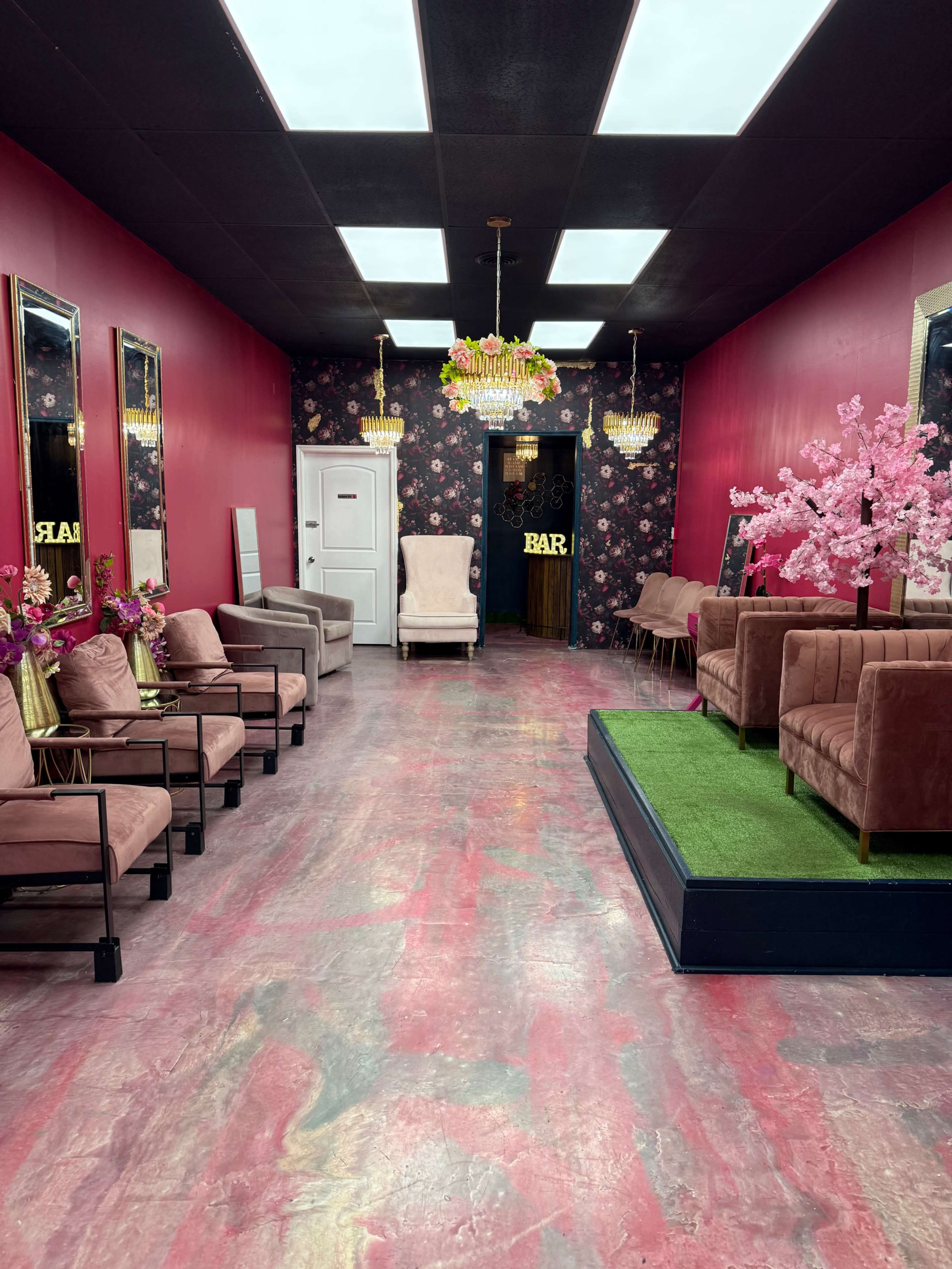 The image shows a brightly colored salon or lounge with pink walls, floral wallpaper, chandeliers, and various seating arrangements, including plush chairs and sofas.