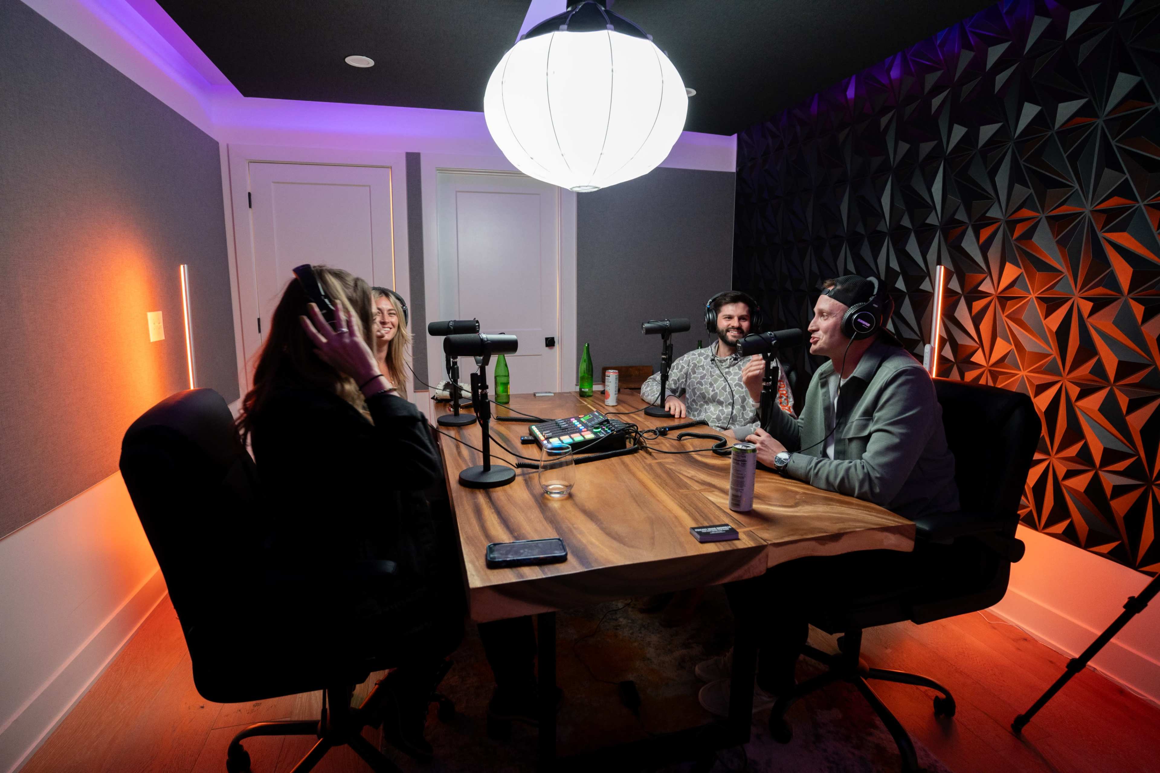 A group of four people sits around a large wooden table in a recording studio, with microphones and recording equipment in front of them.