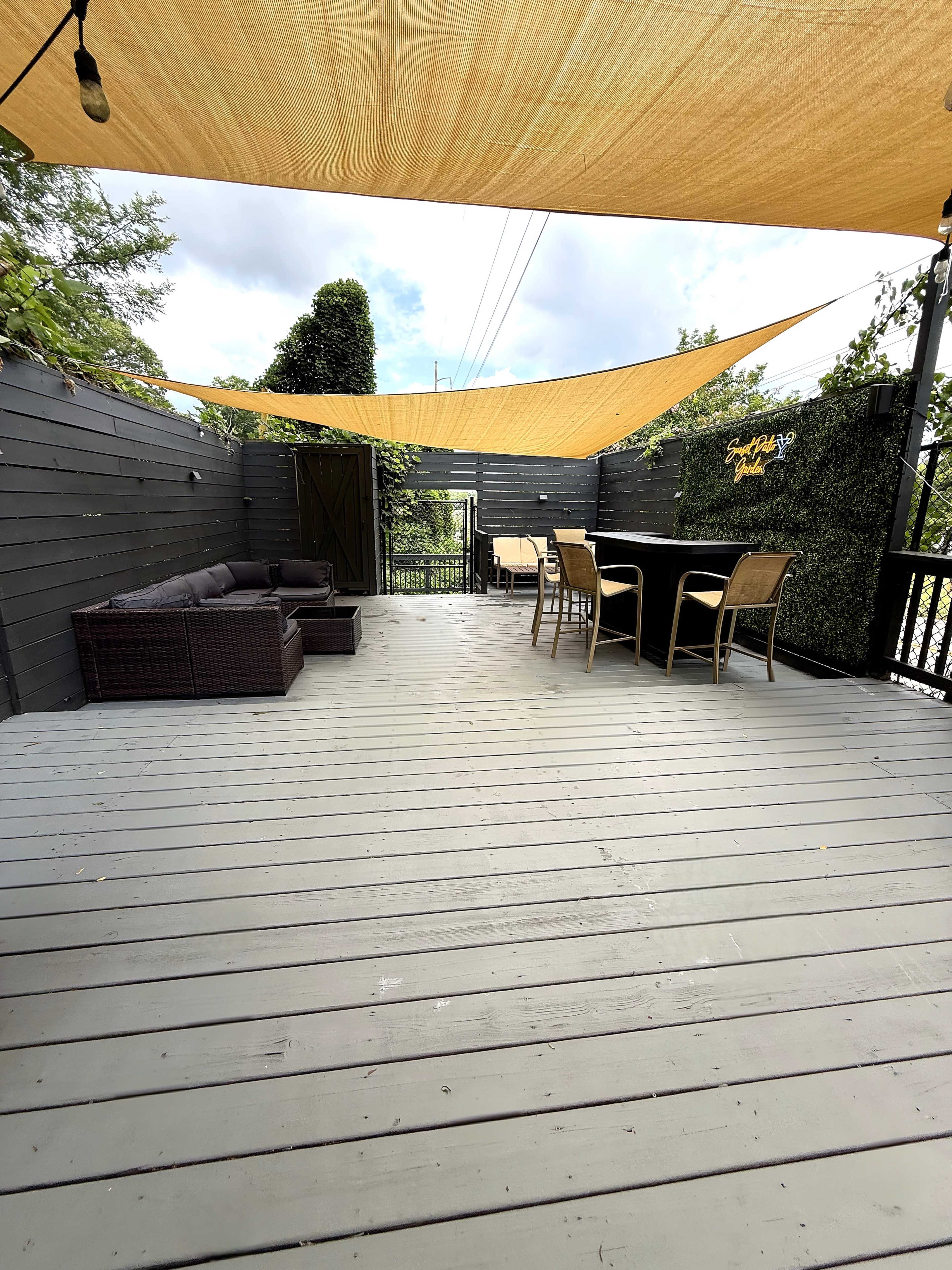 The image shows a spacious outdoor deck with shaded areas provided by yellow canopies, featuring a seating arrangement with a couch and a dining table set.