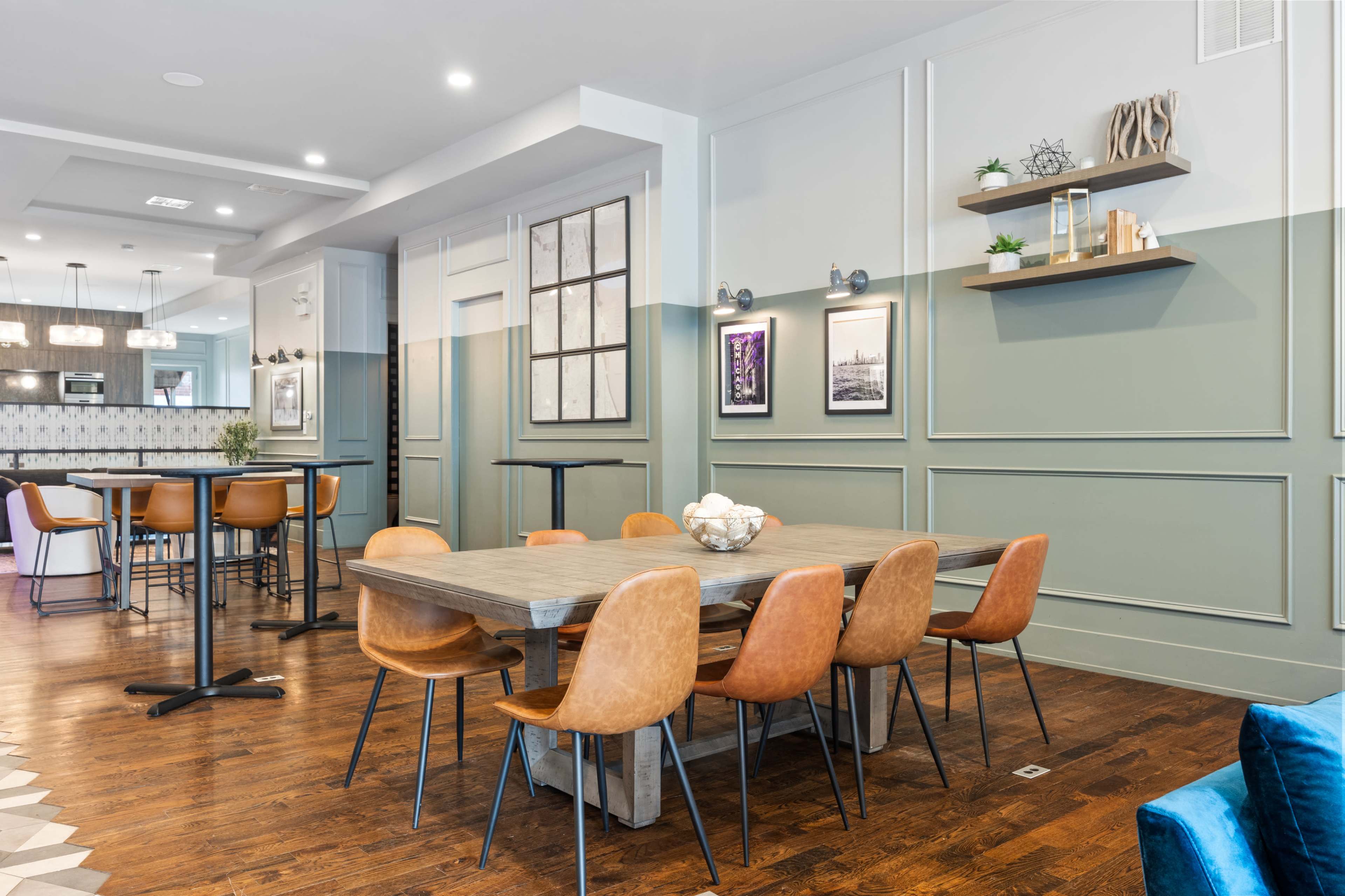A dining area with a long wooden table surrounded by brown and blue chairs, situated in a room with light green and white walls, decorative shelves, and framed artwork.