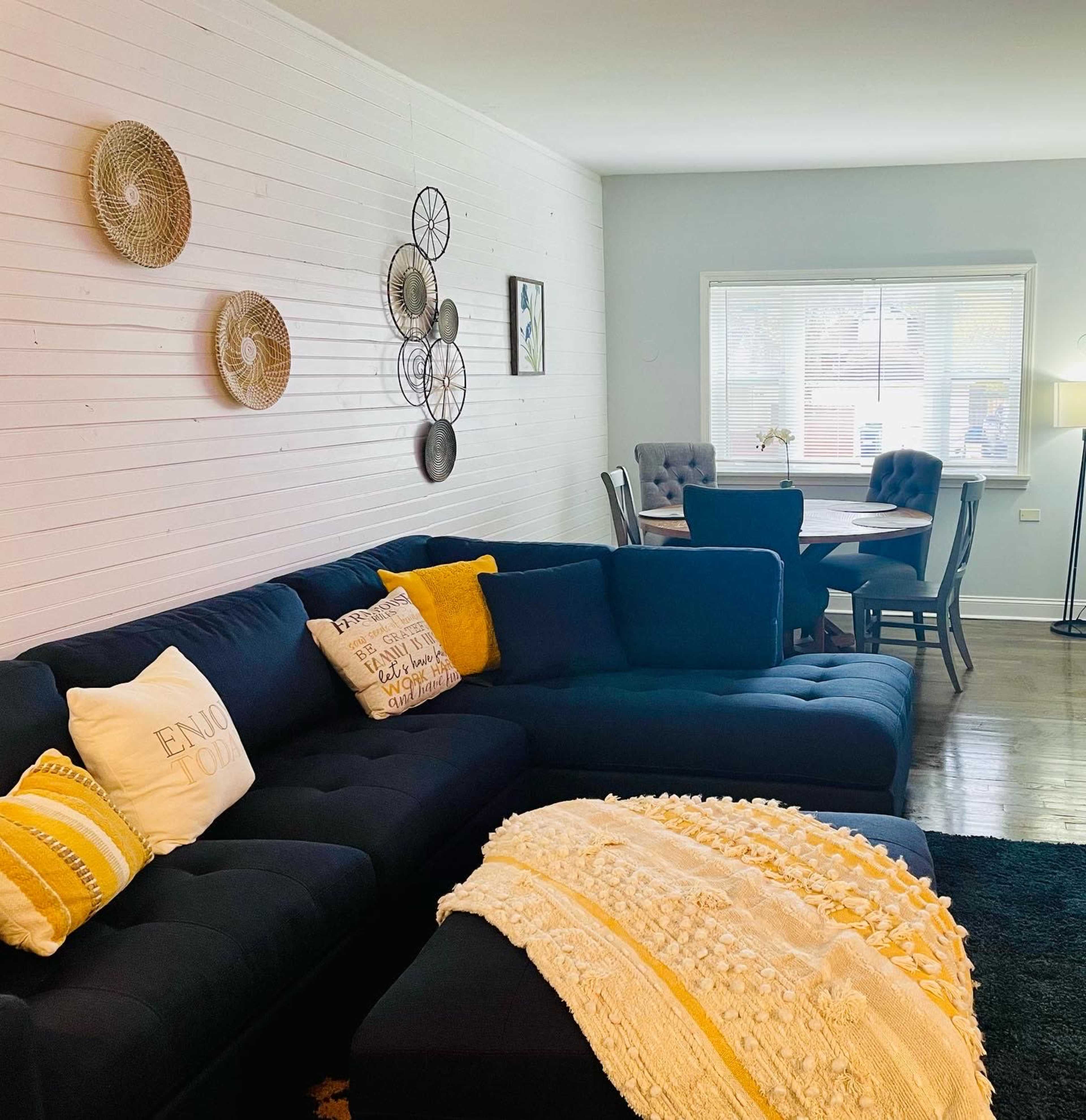 A cozy living room with a dark sectional sofa adorned with decorative pillows, a textured throw blanket, and a dining area featuring a round table and upholstered chairs.
