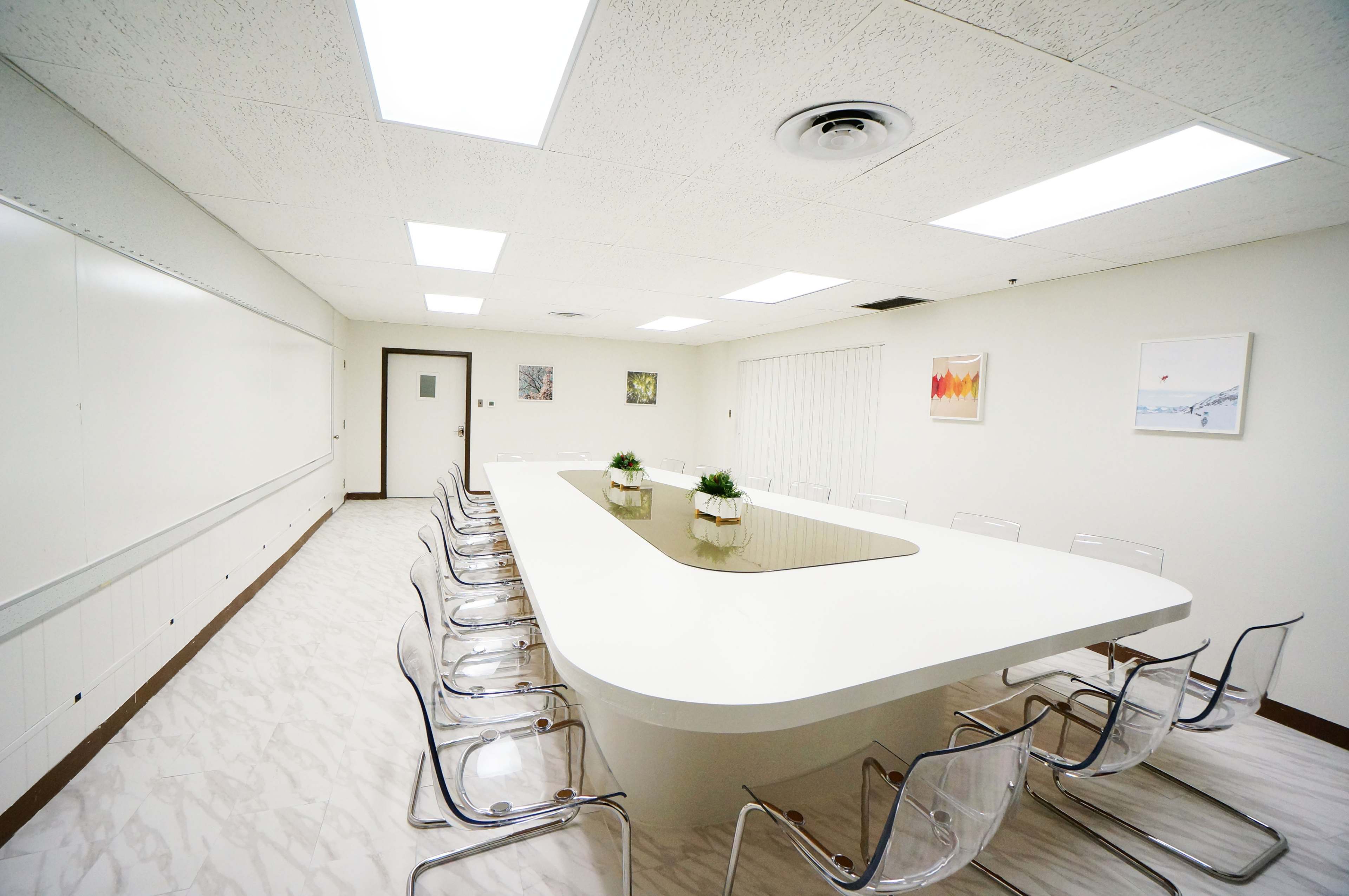 A modern conference room features a large, white oval table surrounded by transparent chairs, with a small plant centerpiece and bright overhead lighting.