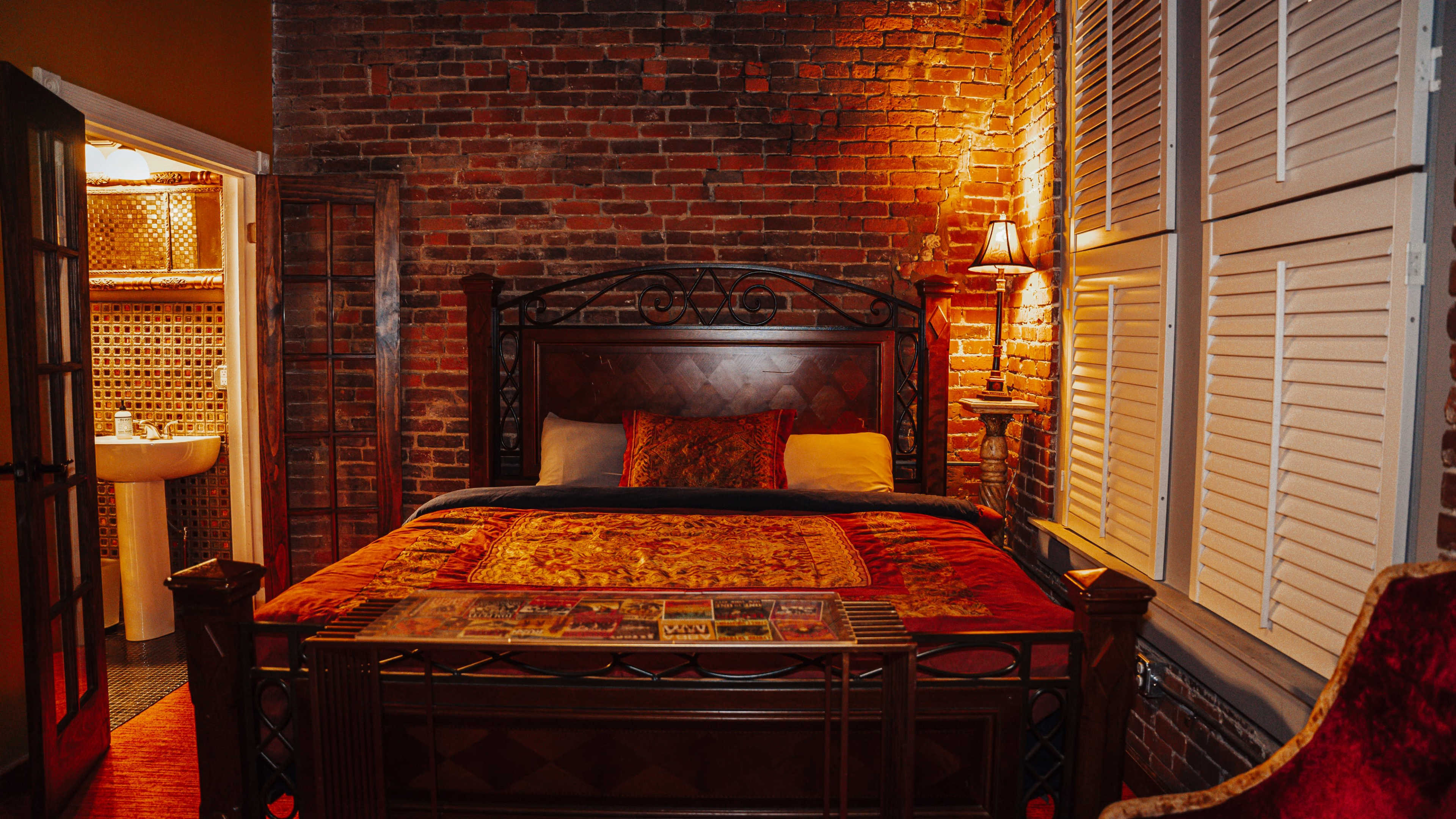 The image shows a warmly lit bedroom featuring a wooden bed with a colorful quilt, exposed brick walls, and a bathroom visible through an open door.