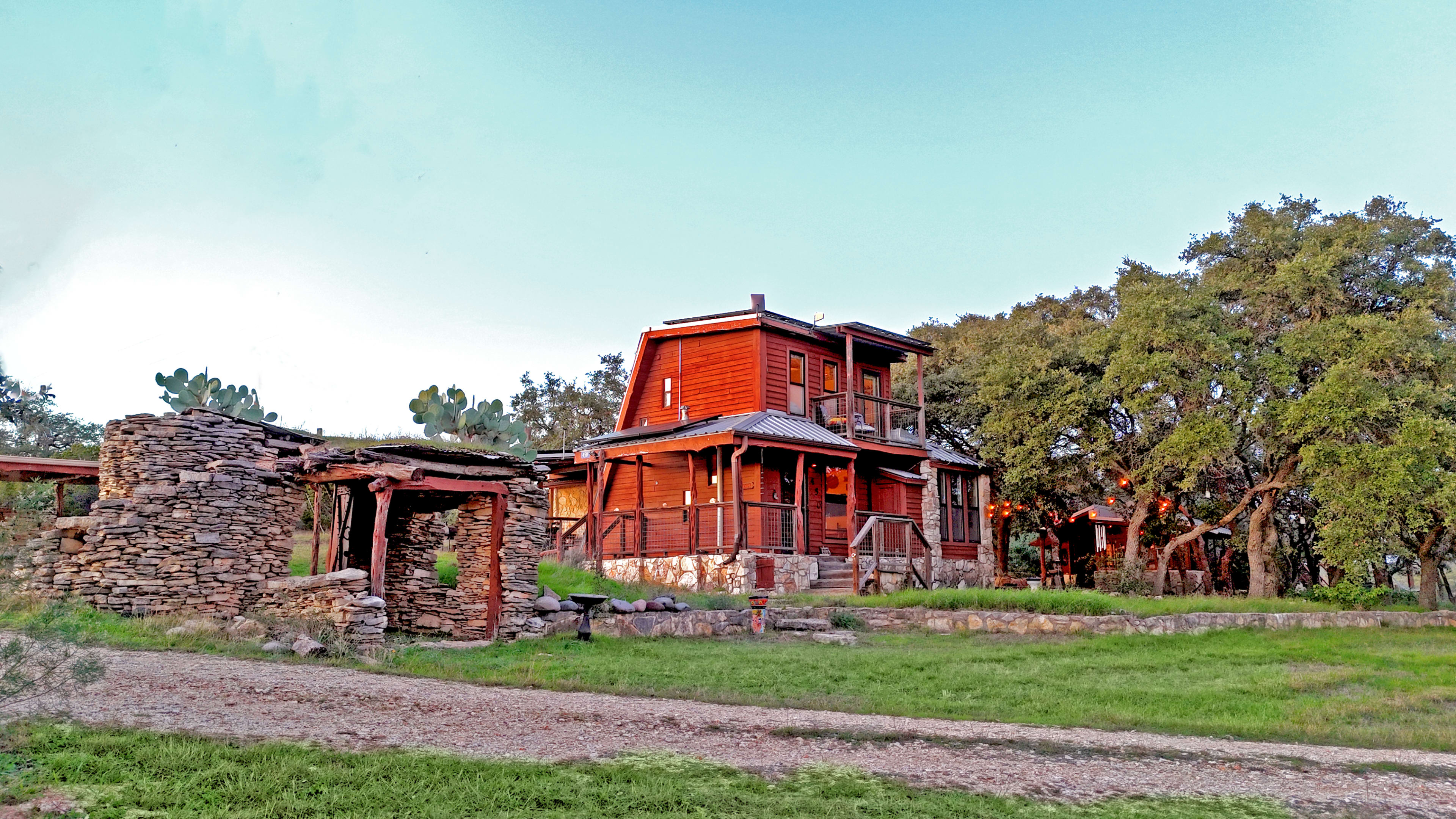 A two-story wooden house with a stone structure nearby is surrounded by grass and trees.