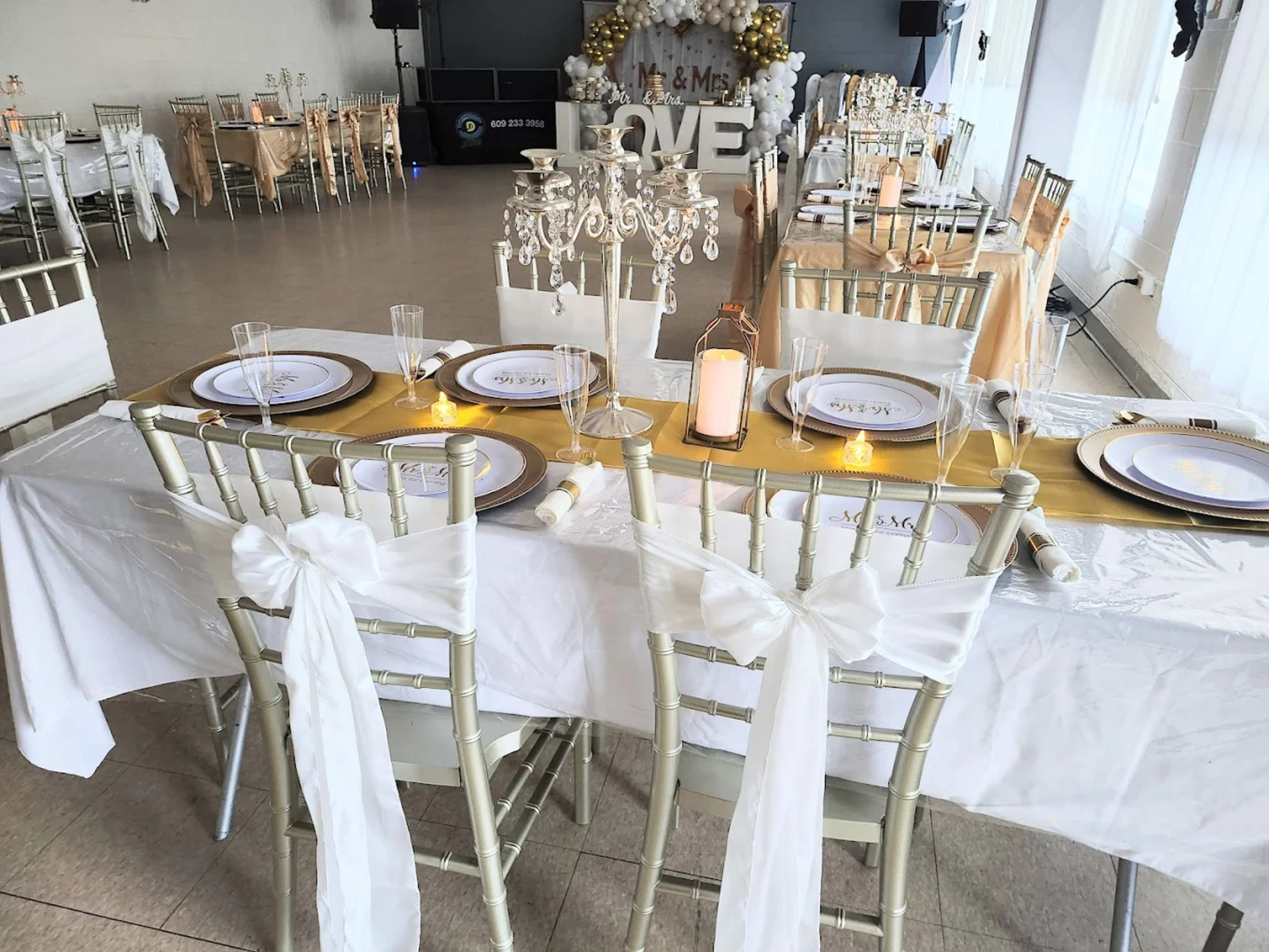 A banquet hall is set up for an event with elegantly arranged tables featuring white tablecloths, gold accents, and decorative centerpieces.