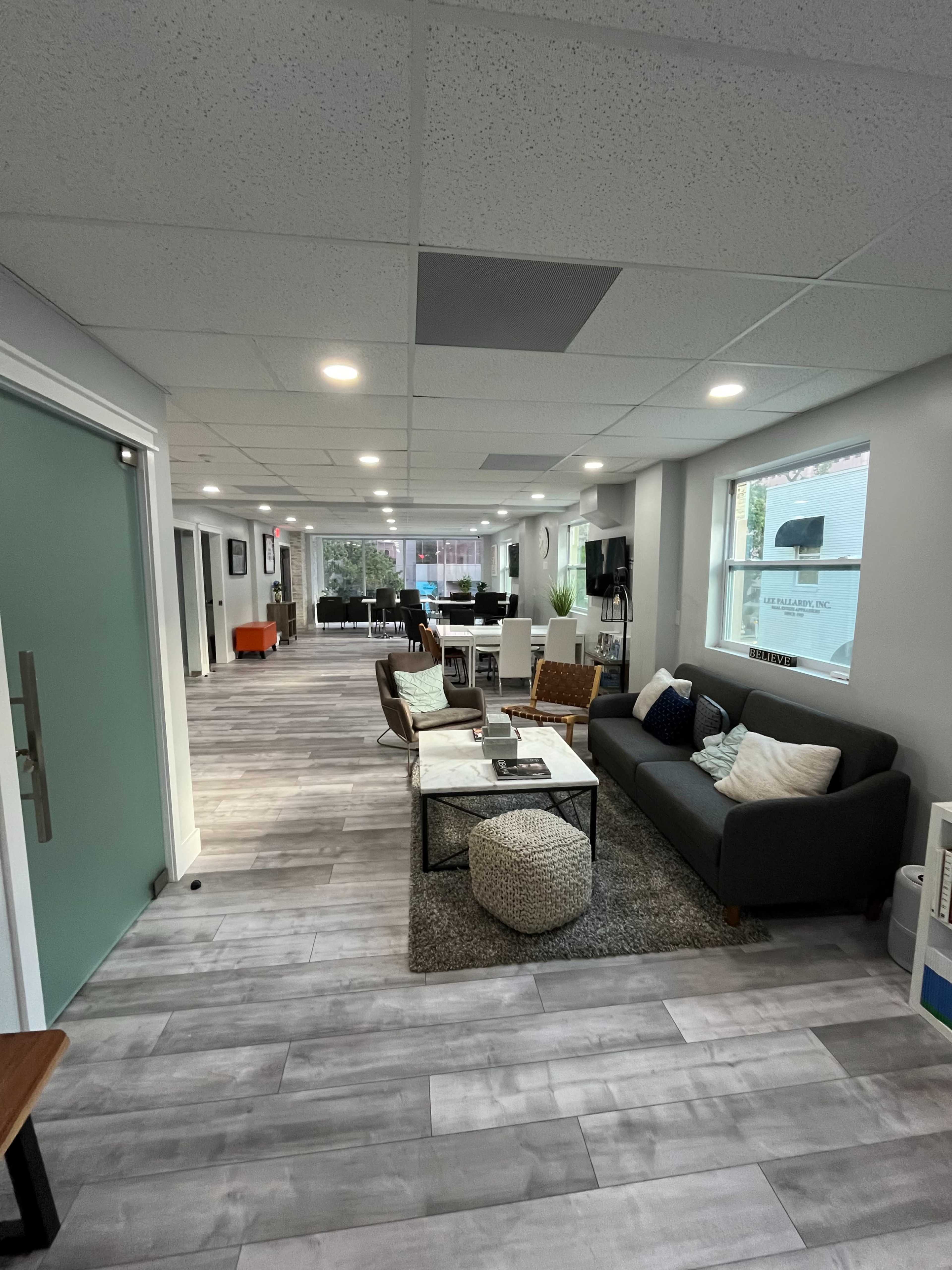 The image shows a modern office interior with a seating area, a light gray tile floor, and large windows along one side.