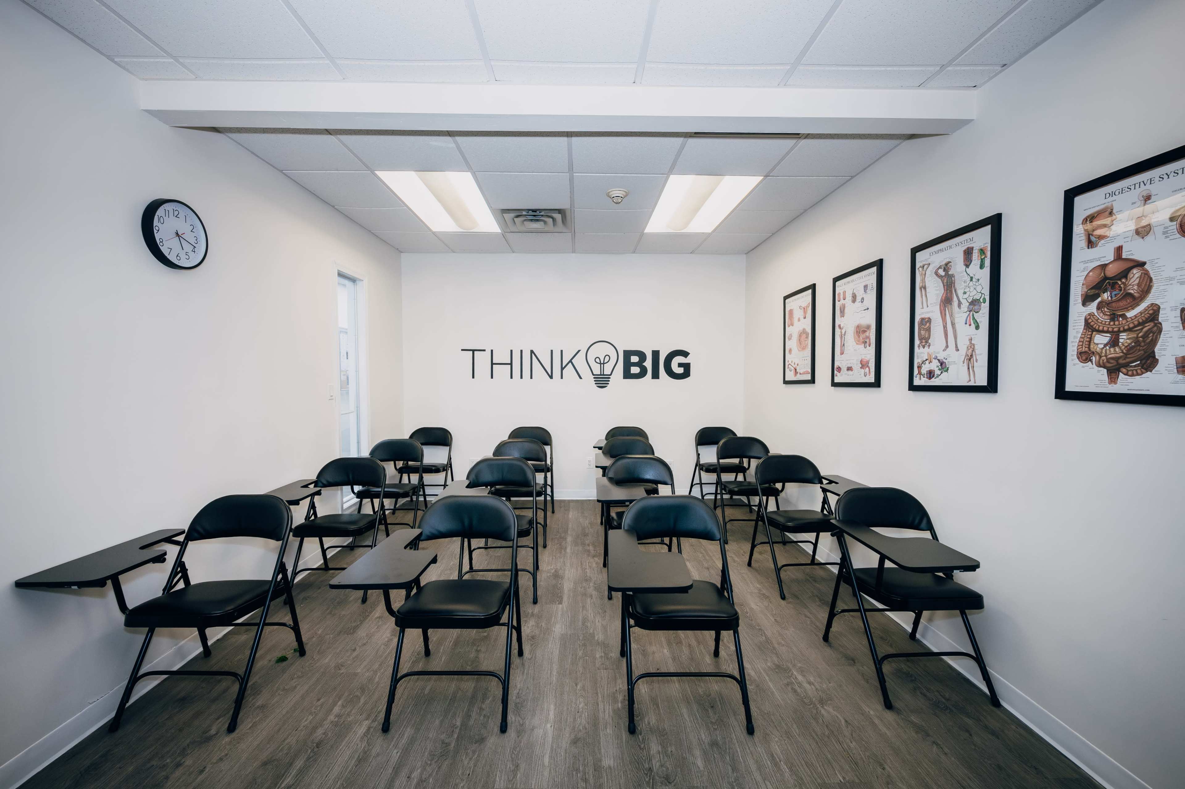 The image shows a classroom with empty black folding chairs arranged in rows facing a wall that displays the phrase "THINK BIG" along with educational posters.
