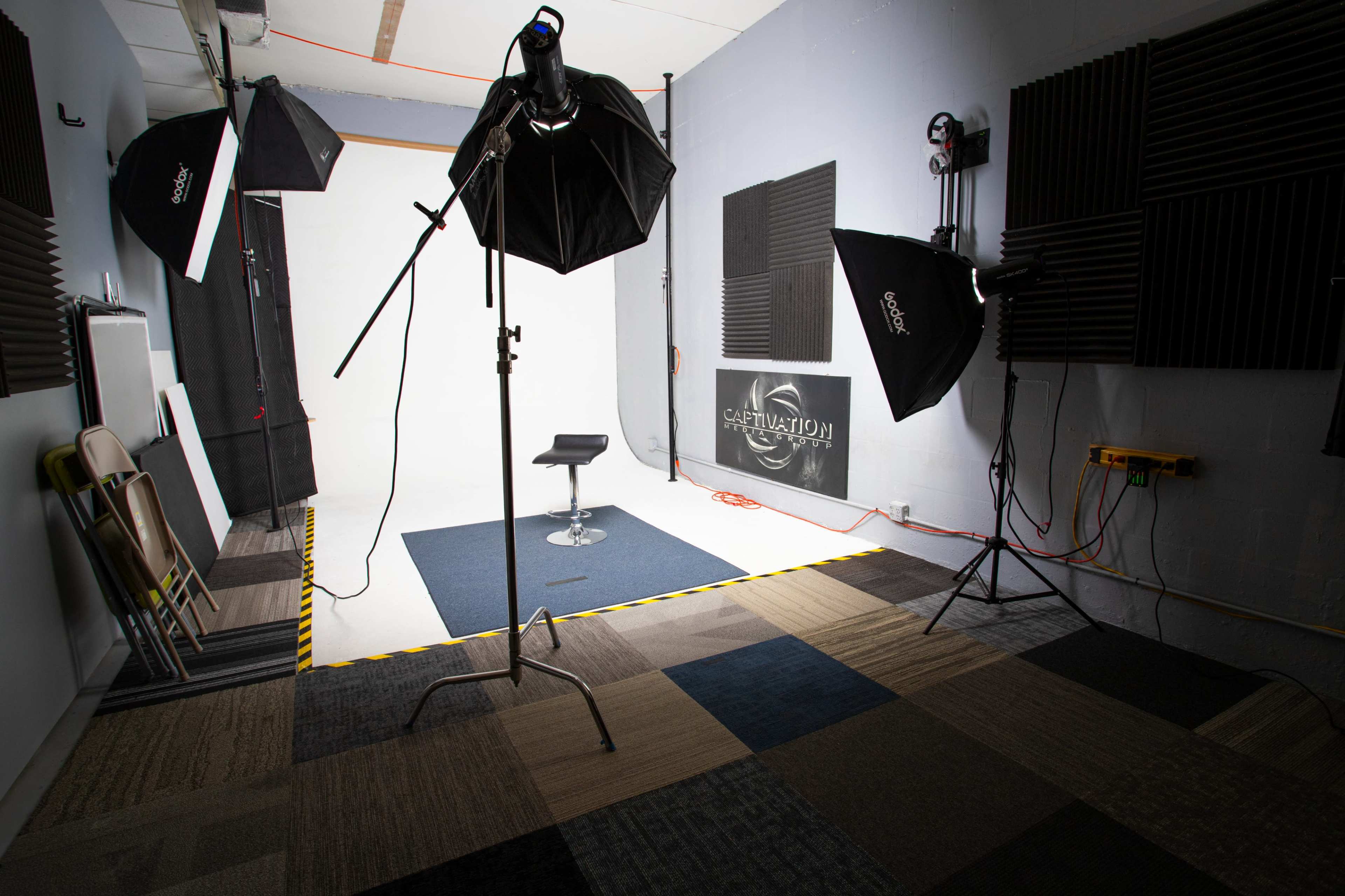 The image shows a photography studio with a central stool, umbrella lights, and a plain backdrop, all set on a patterned floor.