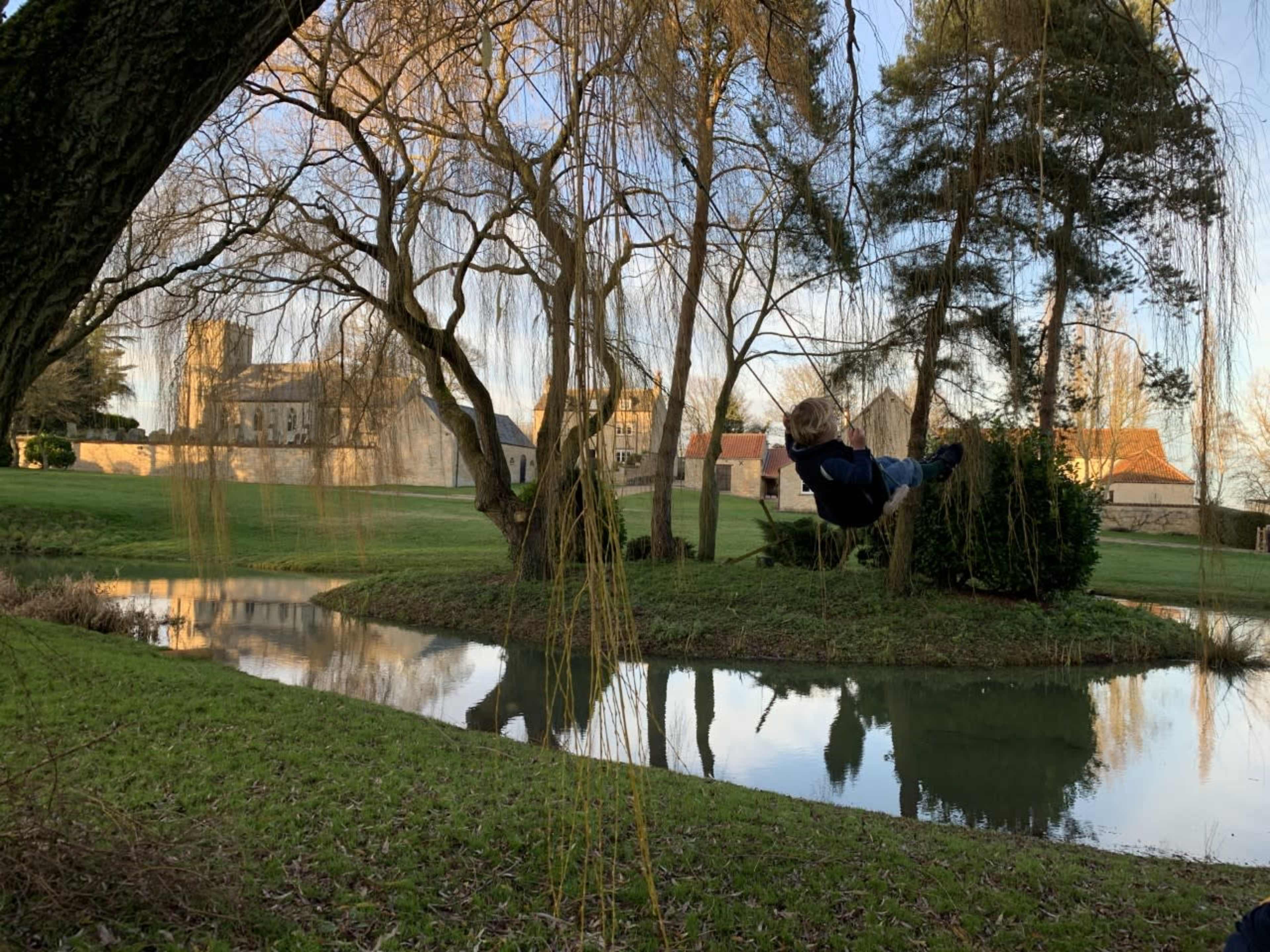 A child swings from a tree near a serene pond, with a historic building visible in the background.