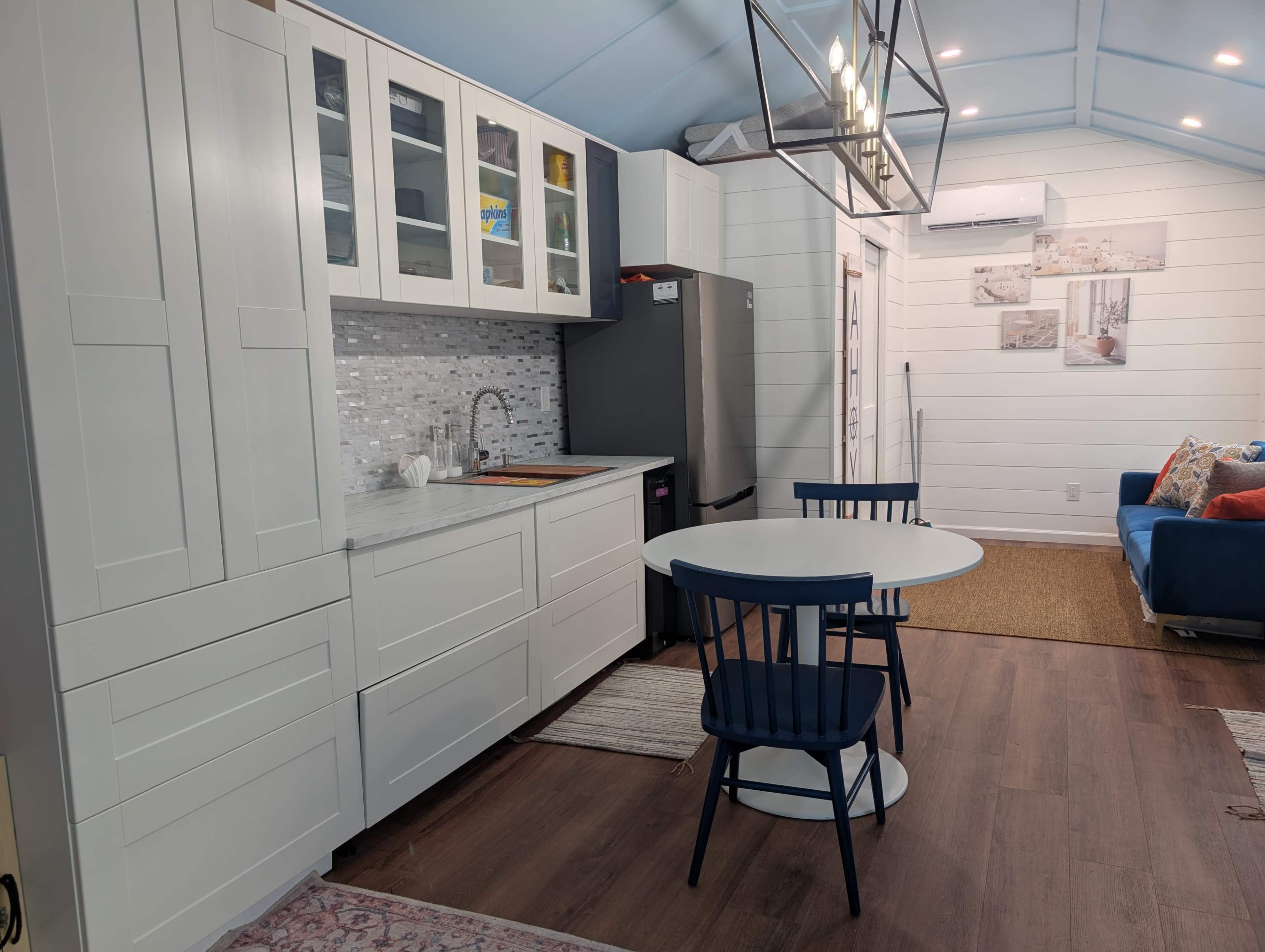 The image shows a compact kitchen area with white cabinetry, a stainless steel refrigerator, and a round dining table with blue chairs, located in a well-lit, modern space.