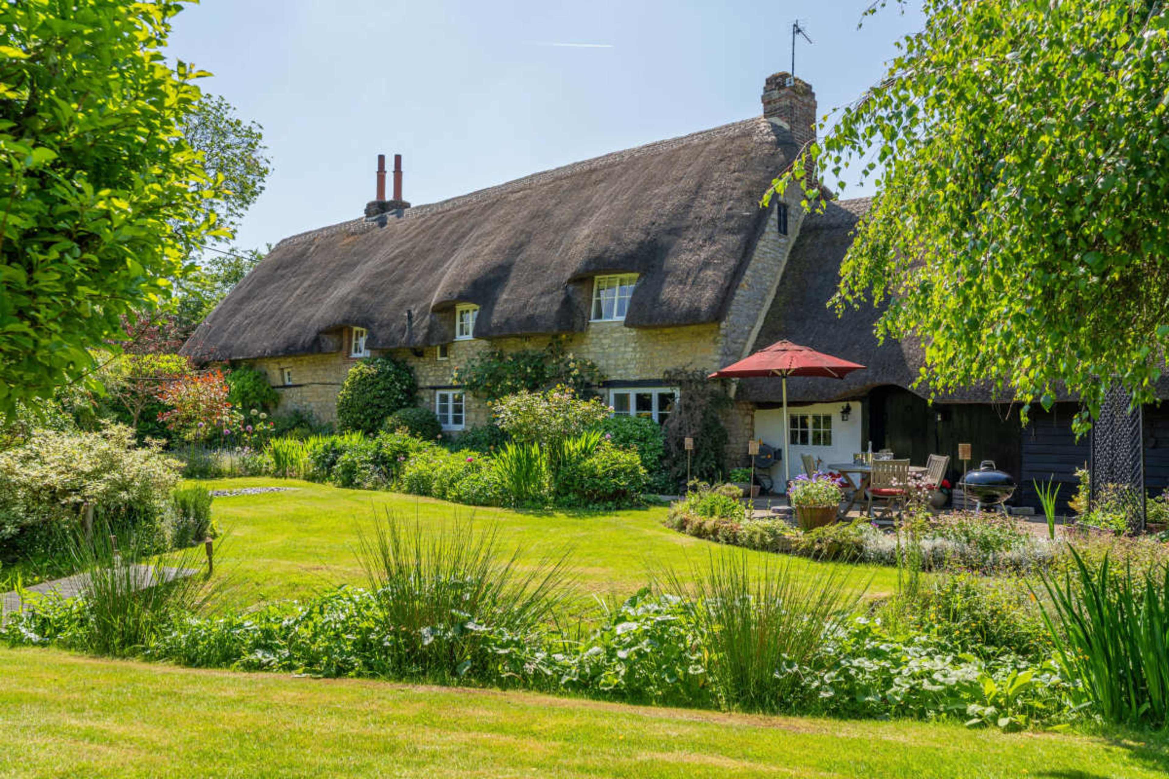 A thatched cottage with a manicured garden and patio area is surrounded by lush greenery and flowering plants.