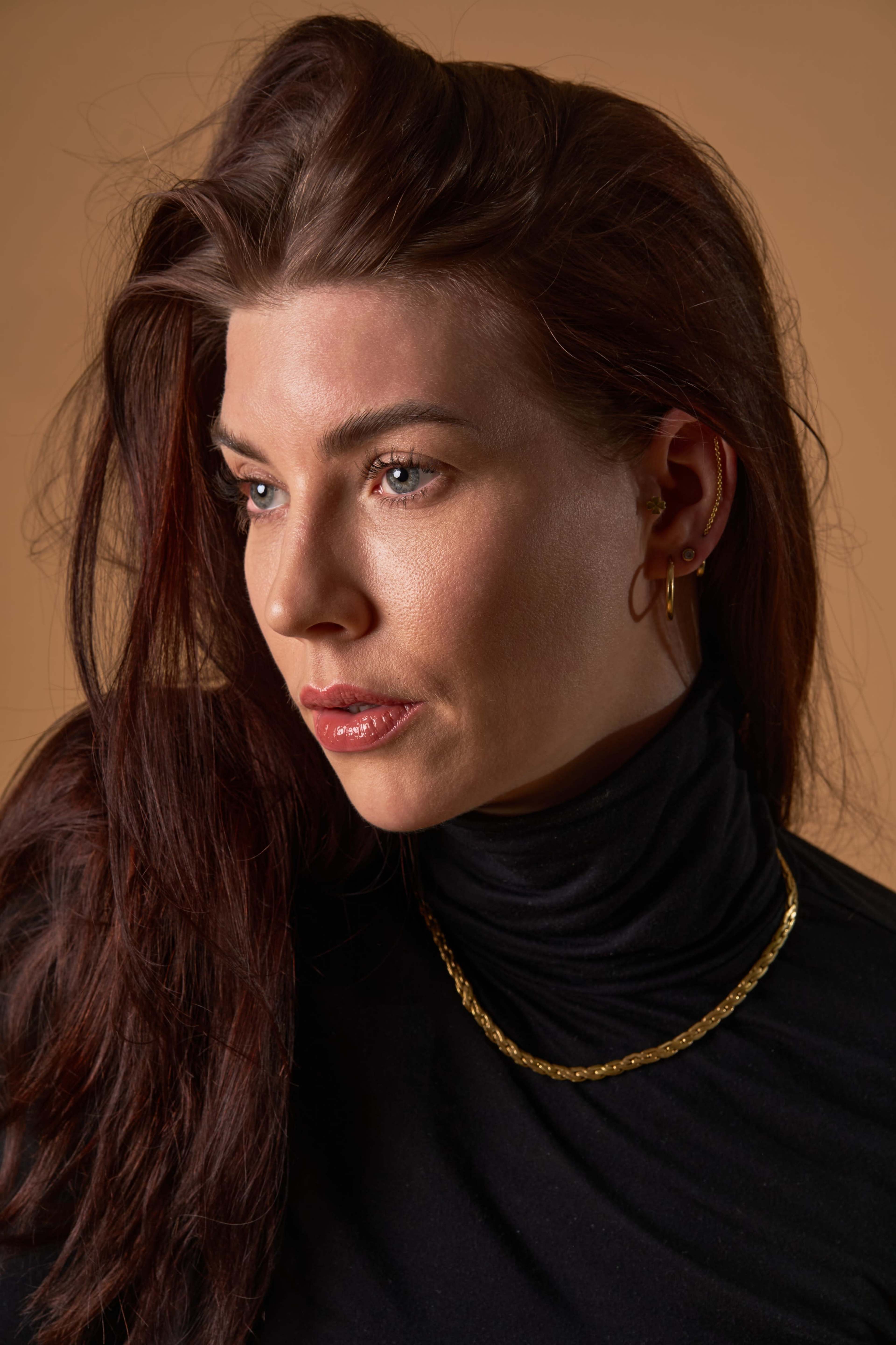 A woman with long, dark hair poses against a beige background while wearing a black turtleneck and gold jewelry.