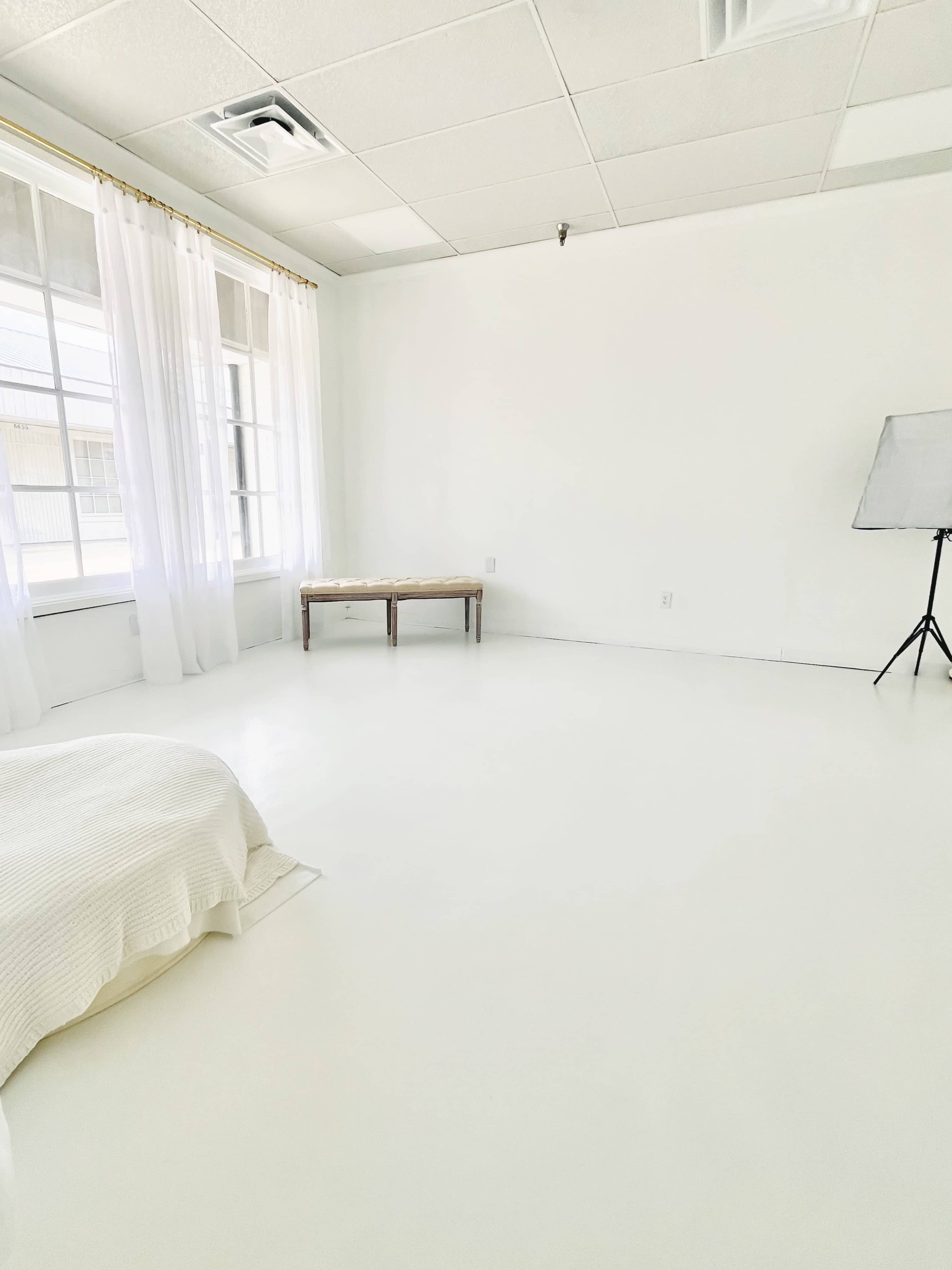 The image shows a bright, minimalistic room with white walls and flooring, featuring a bed with a light-colored blanket and a wooden bench near a large window with sheer curtains.