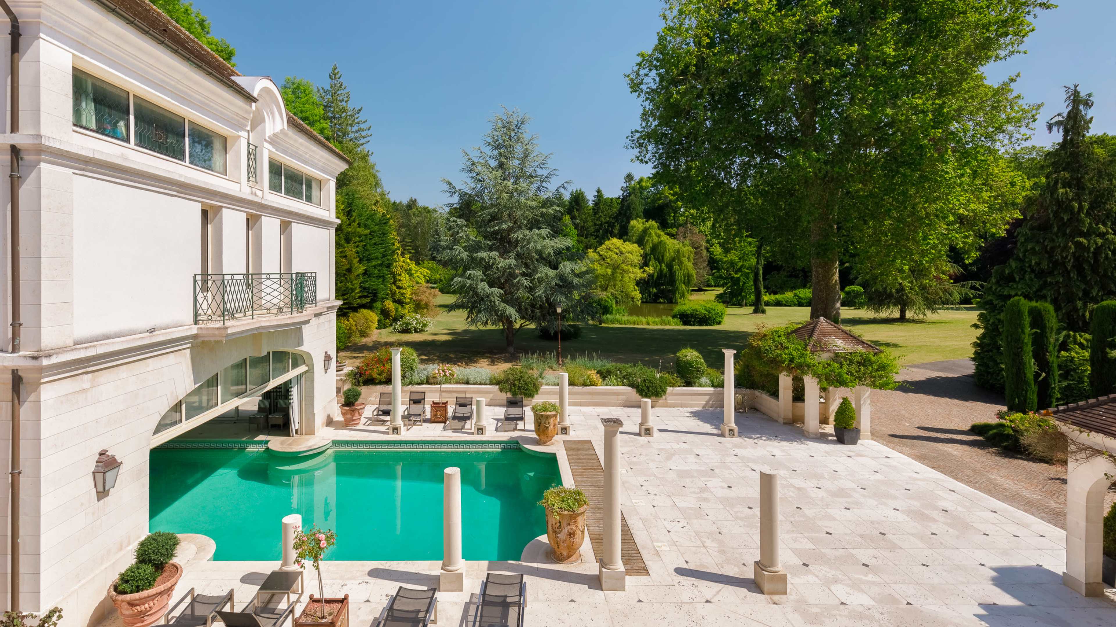 A large swimming pool with stone columns is situated in an outdoor area beside a white villa, surrounded by lush greenery and trees.