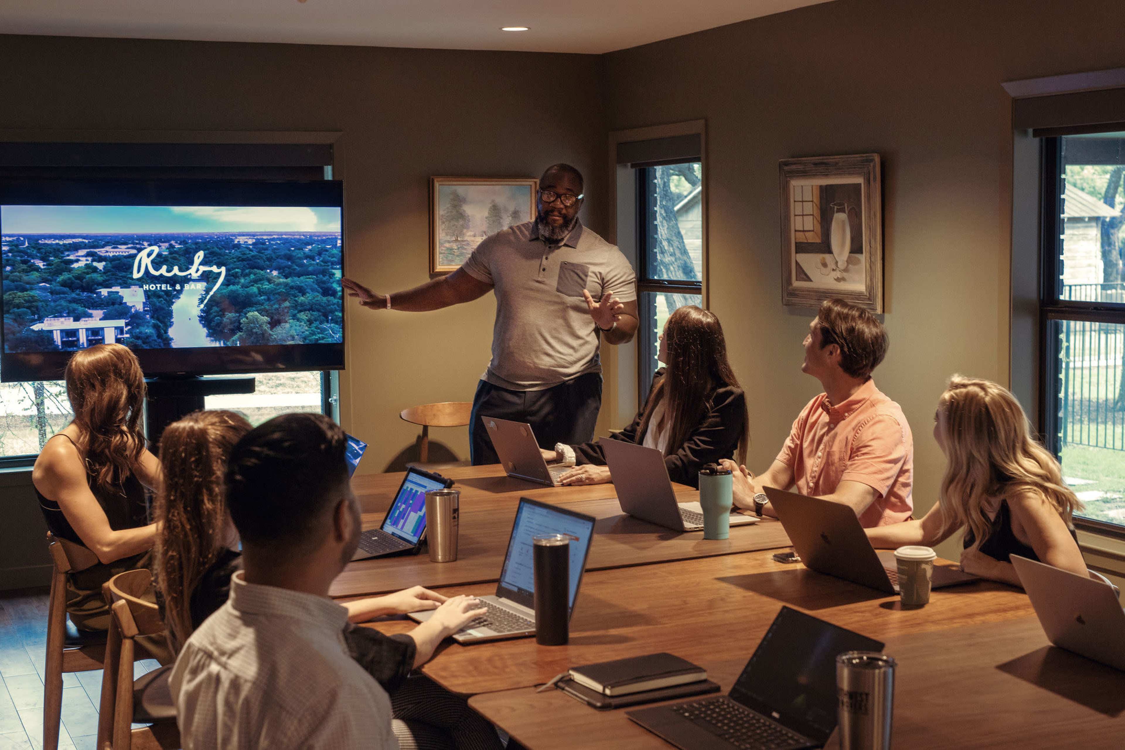 A diverse group of people is seated around a table in a conference room, listening to a presenter who is showing a slideshow on a large screen.
