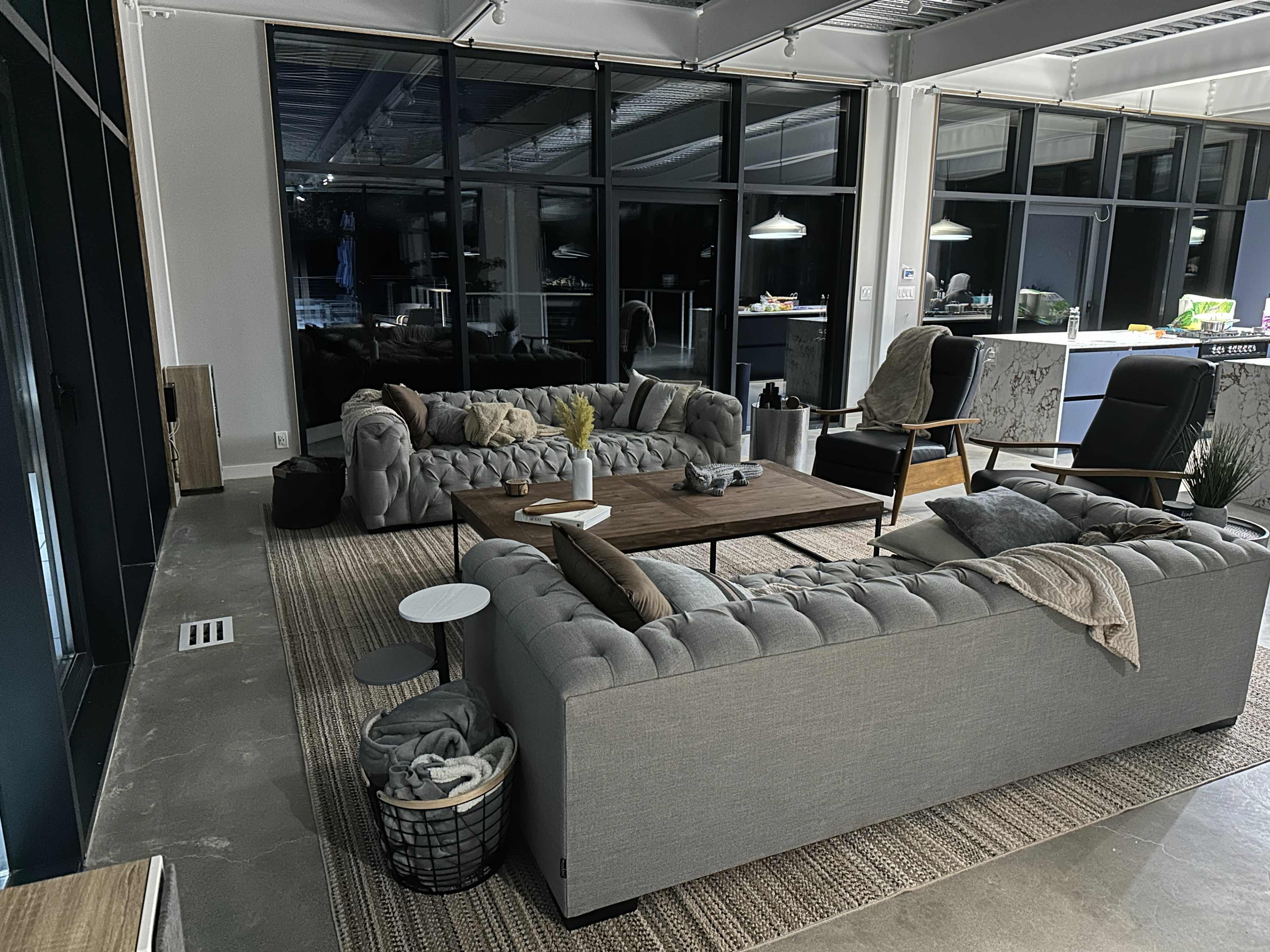 A modern living room with two grey tufted sofas, a wooden coffee table, and large windows allowing ample light in.