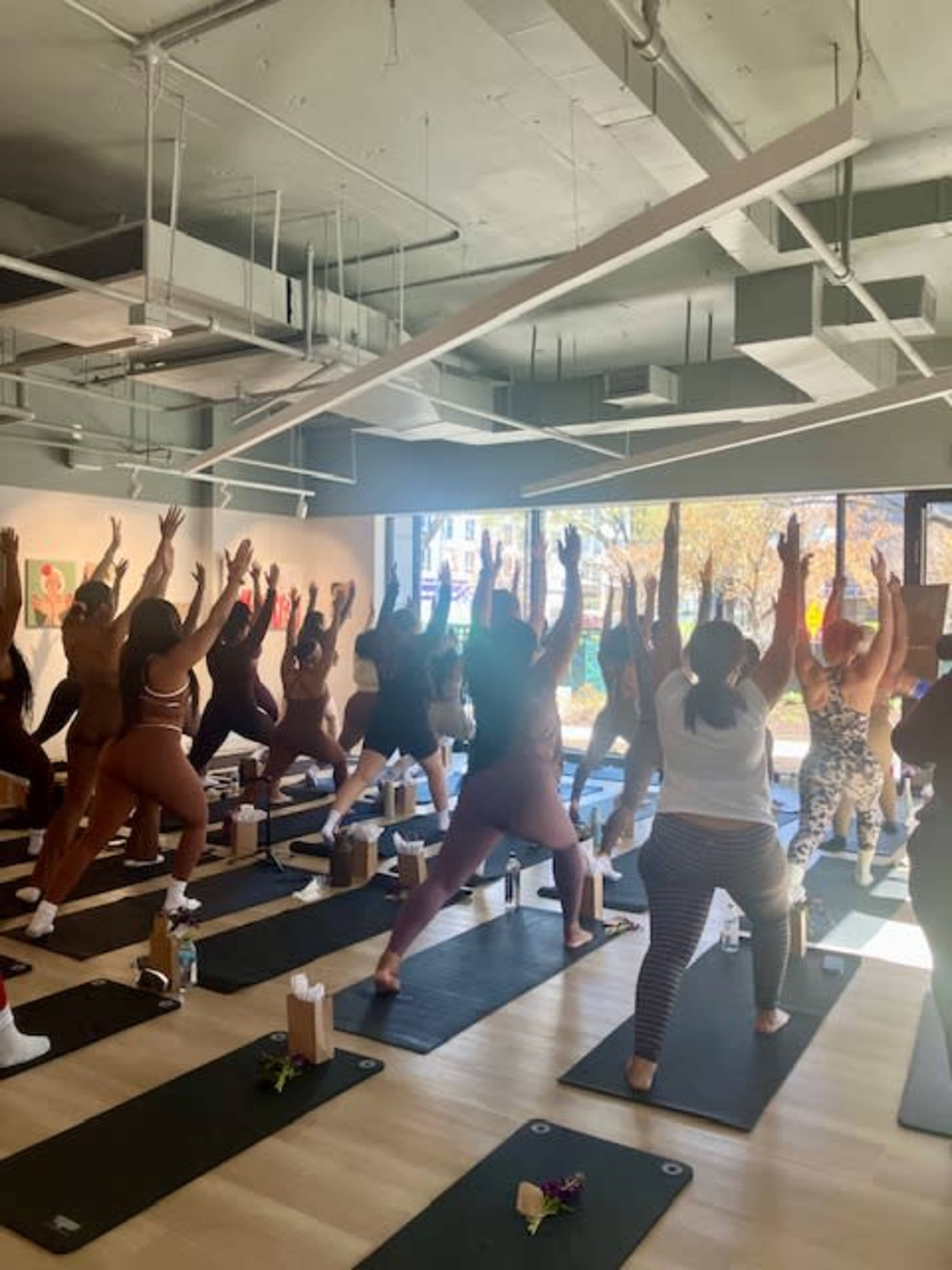 A group of individuals in workout attire practice yoga in a brightly lit studio with large windows.