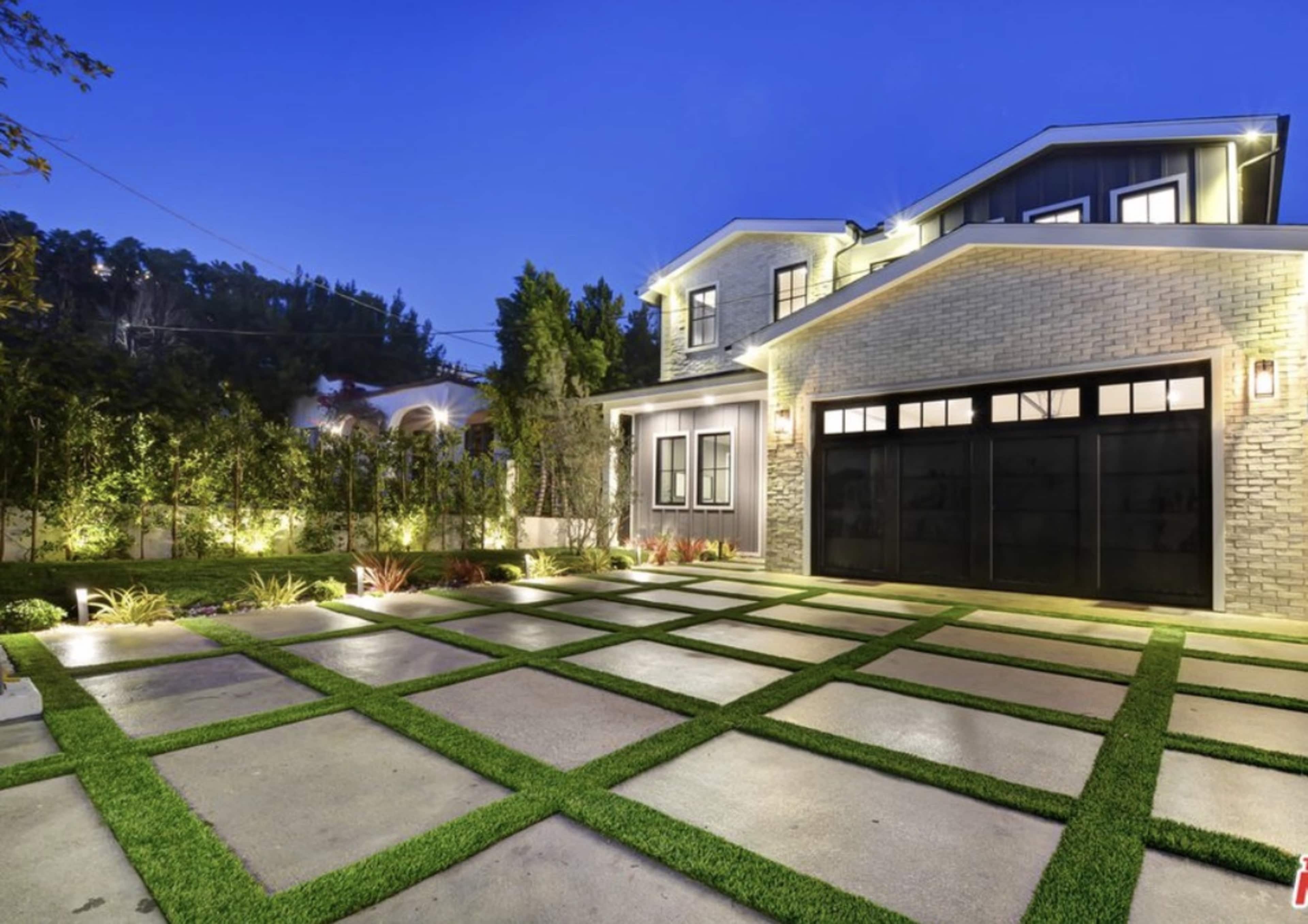 The image shows a modern house with a well-lit facade, featuring a manicured lawn and a geometric concrete driveway with grass strips.