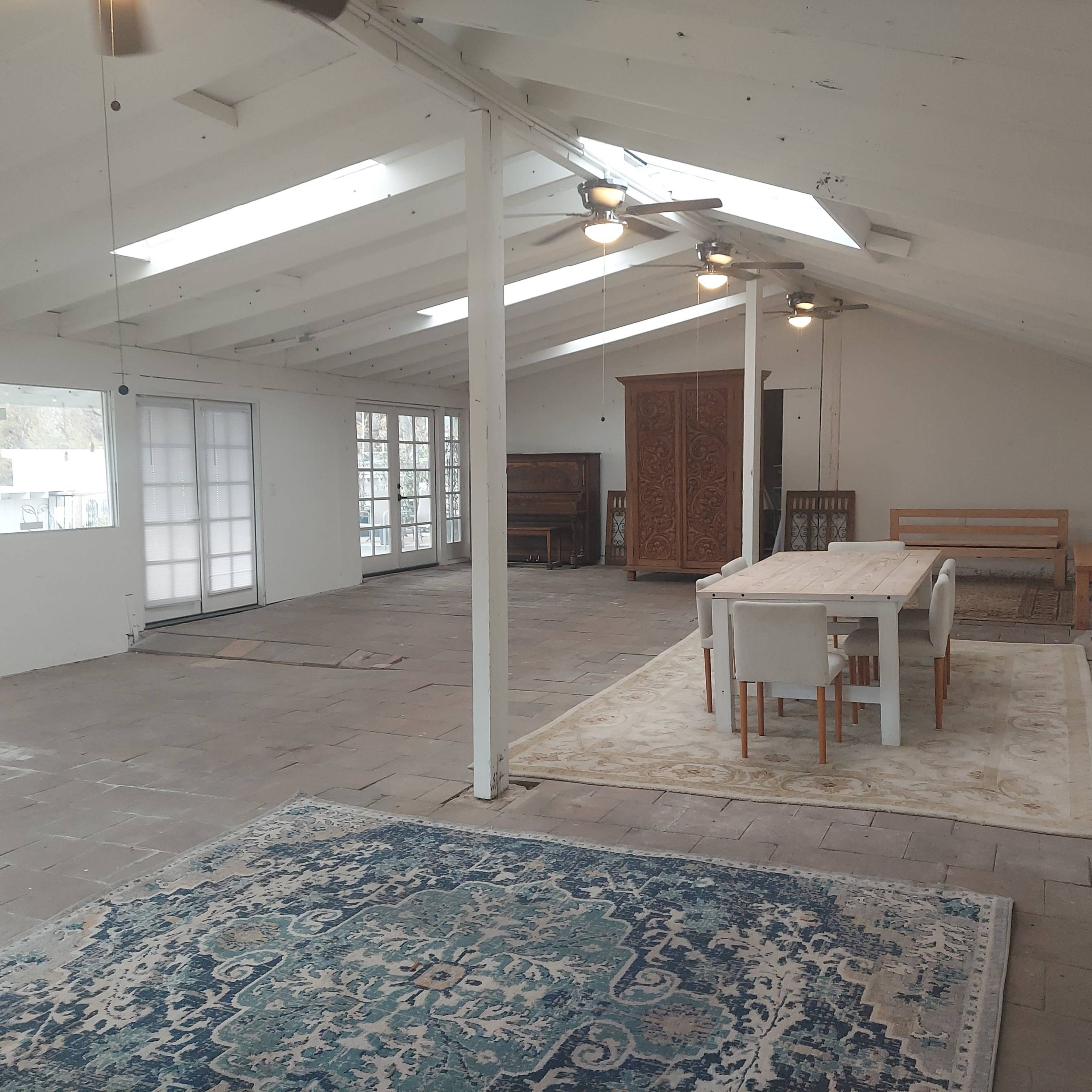 The image shows a spacious, light-filled interior with a dining table, chairs, and a wooden cabinet against a wall.