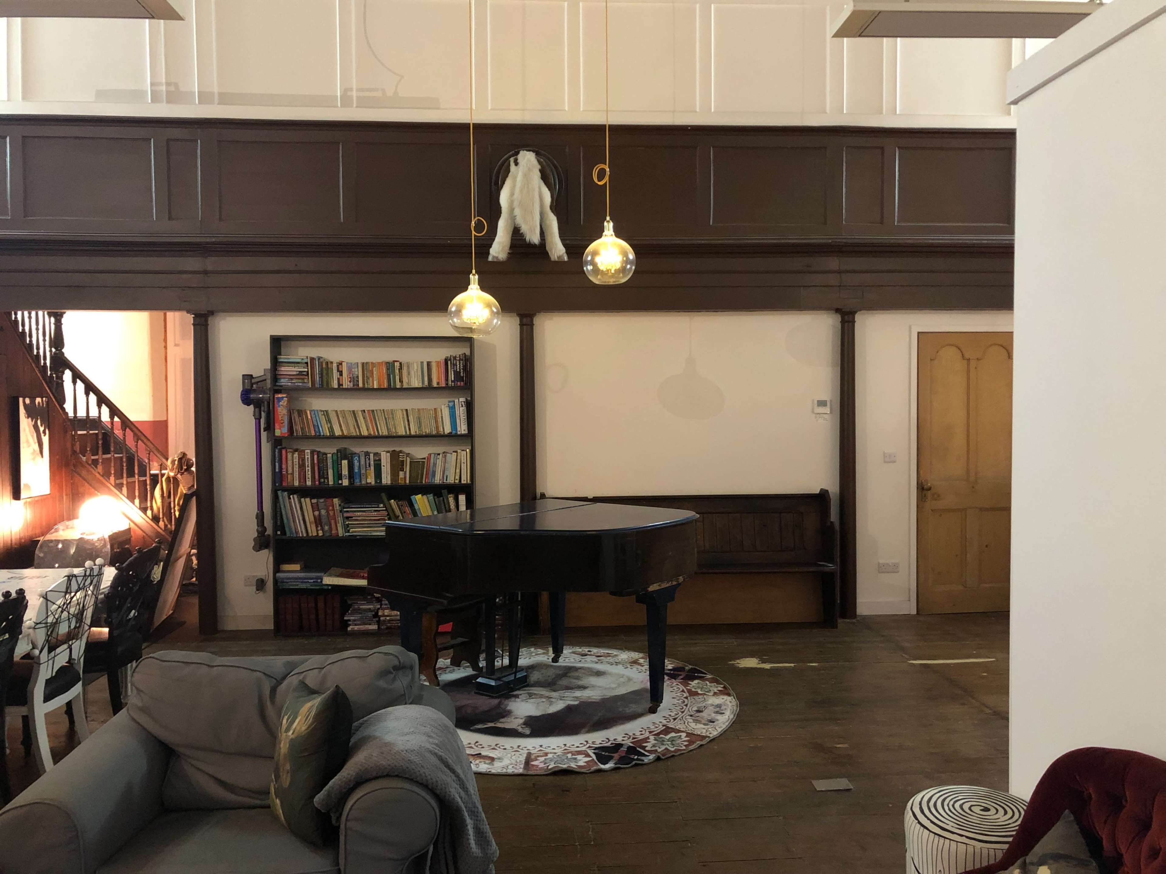 A grand piano sits in the center of a room with a wooden bookcase and a staircase in the background.