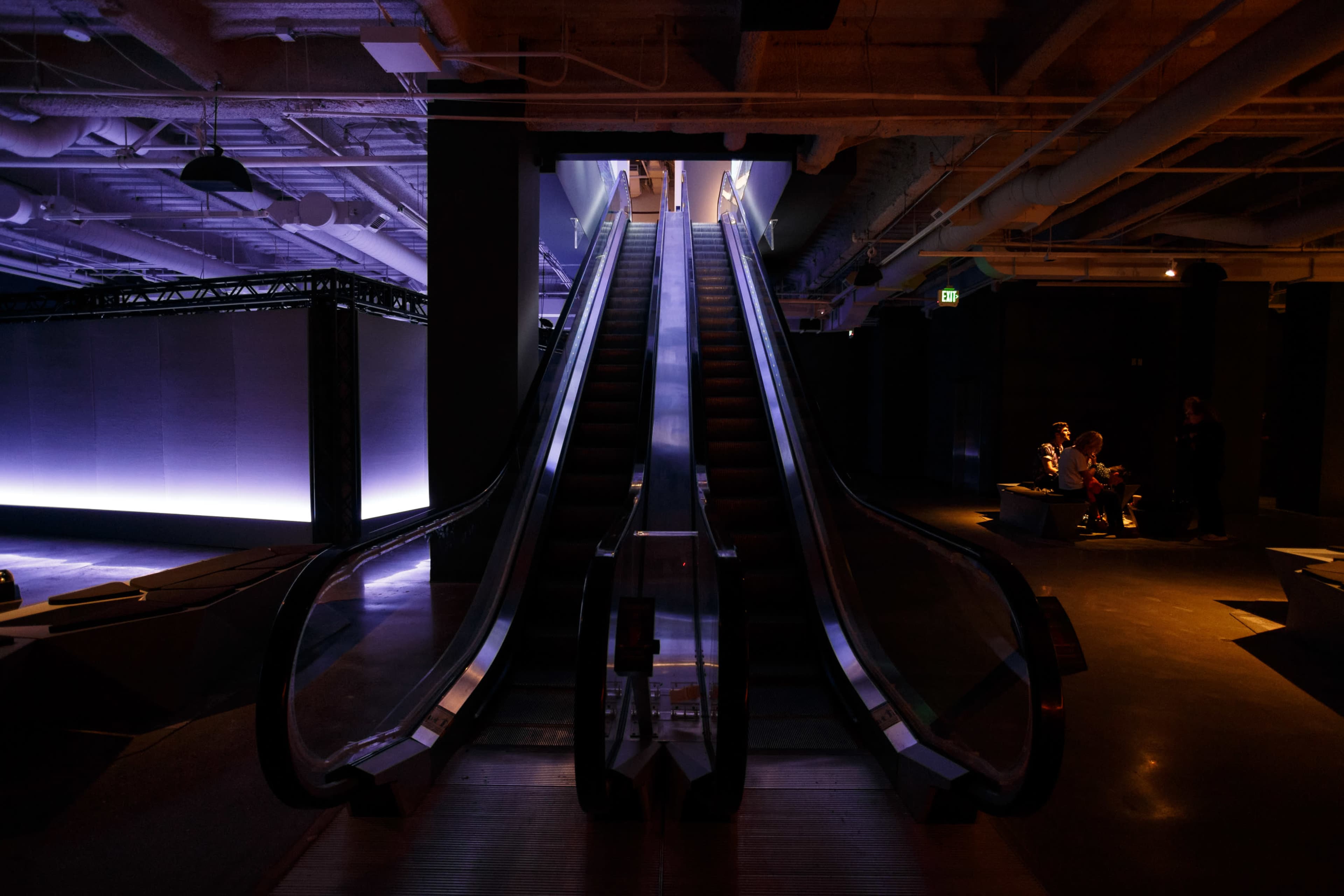 An escalator leading upward in a dimly lit space, with people seated in the background.