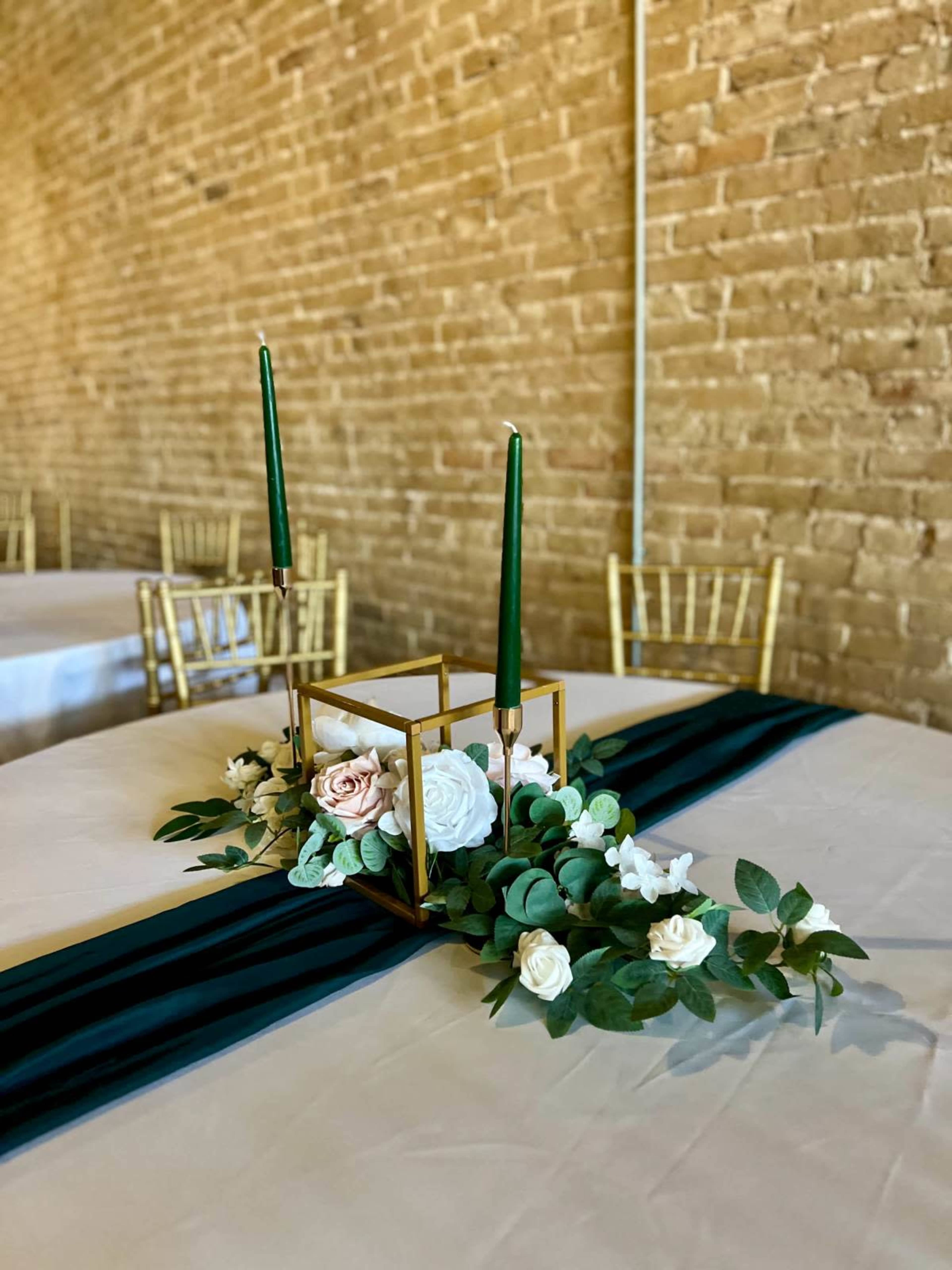 A table set with a greenery and floral centerpiece, featuring two green candlesticks, rests on a white tablecloth in a brick-walled venue.