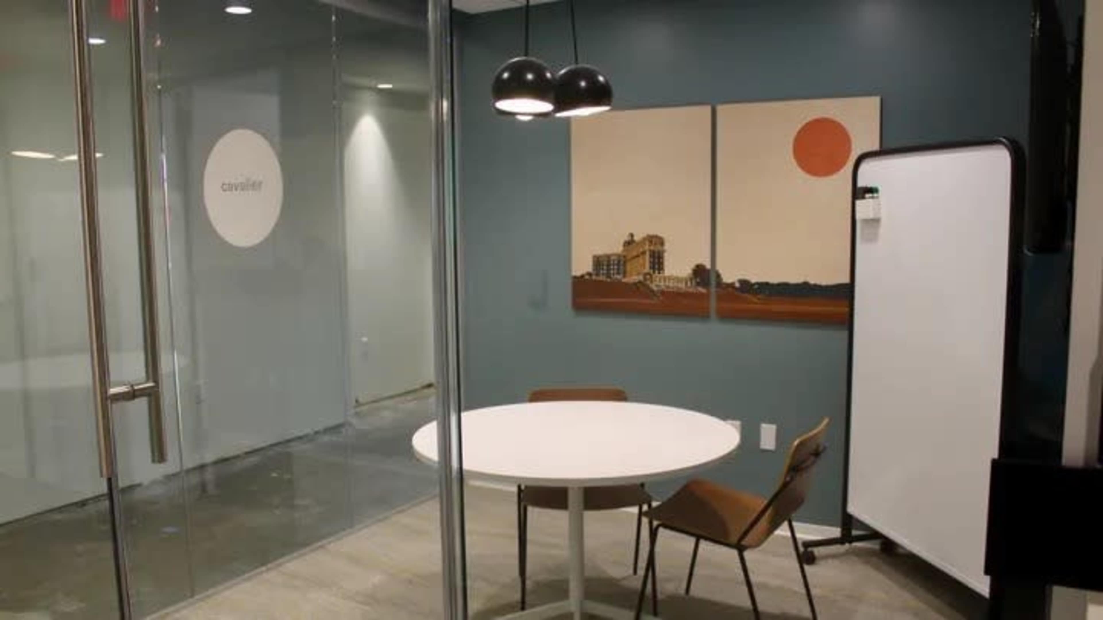 A modern office meeting room with a round table, two chairs, and a large glass wall featuring artwork and a whiteboard.