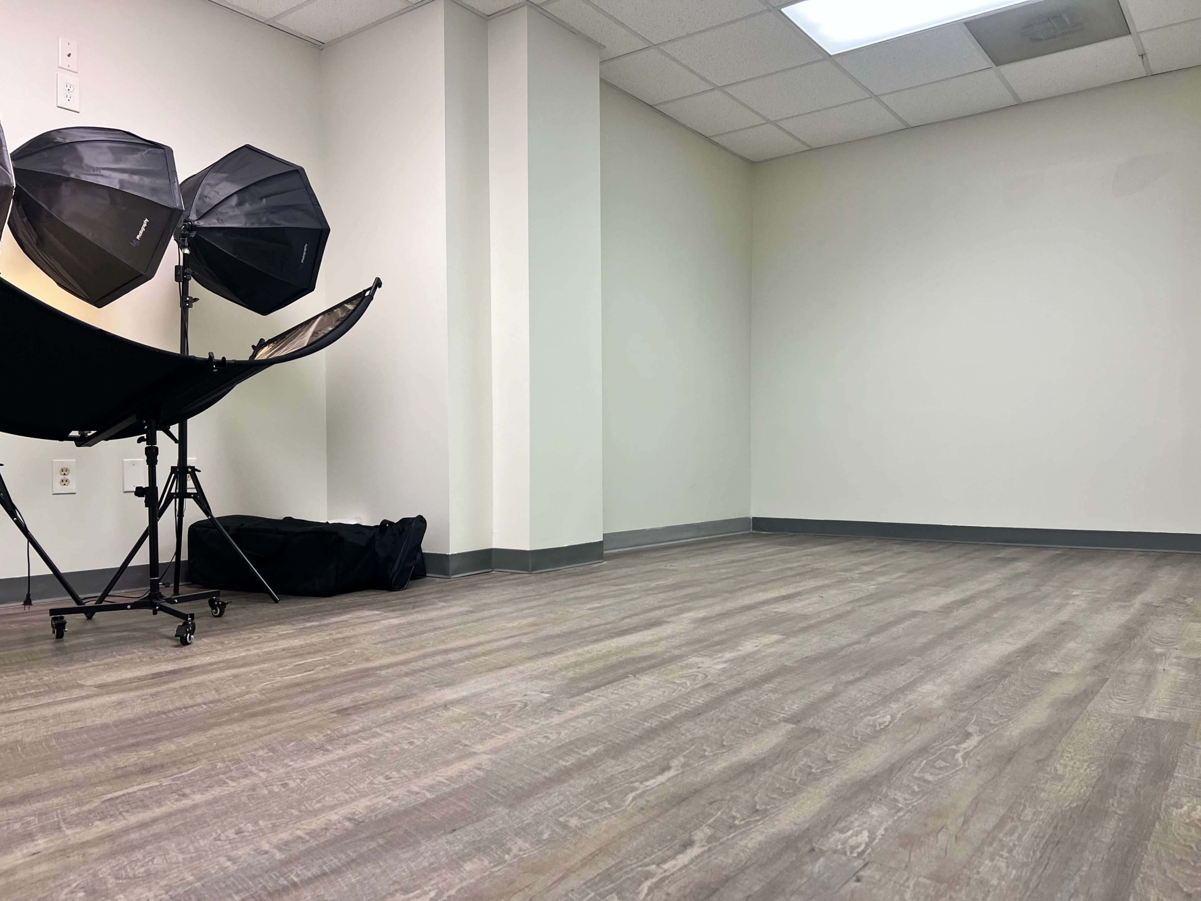 The image shows an empty room with light-colored walls and a wooden floor, featuring two studio lighting equipment setups on the left side.