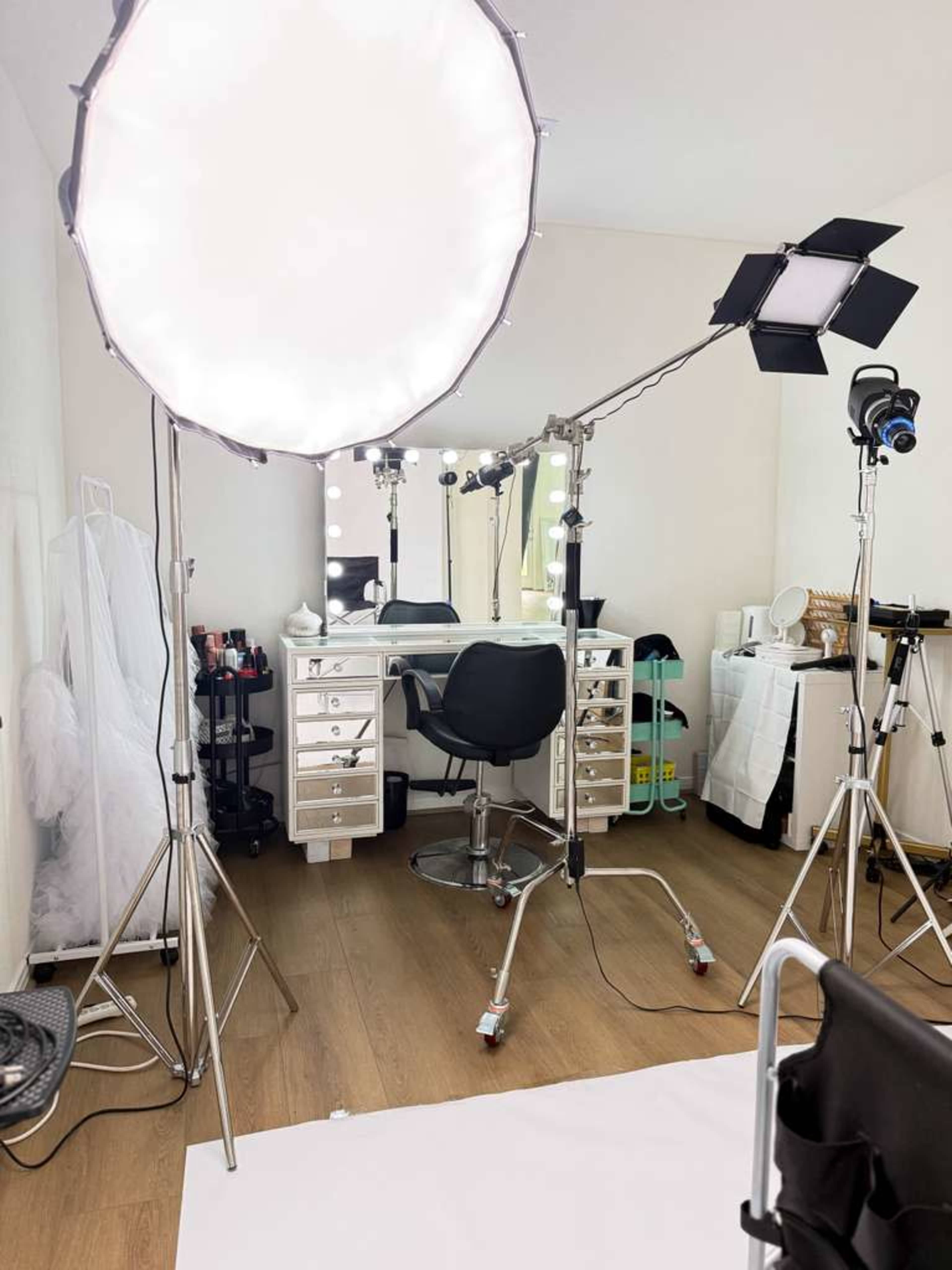 The image shows a well-lit makeup studio with a vanity area, equipped with multiple light sources and a rolling chair on a wooden floor.