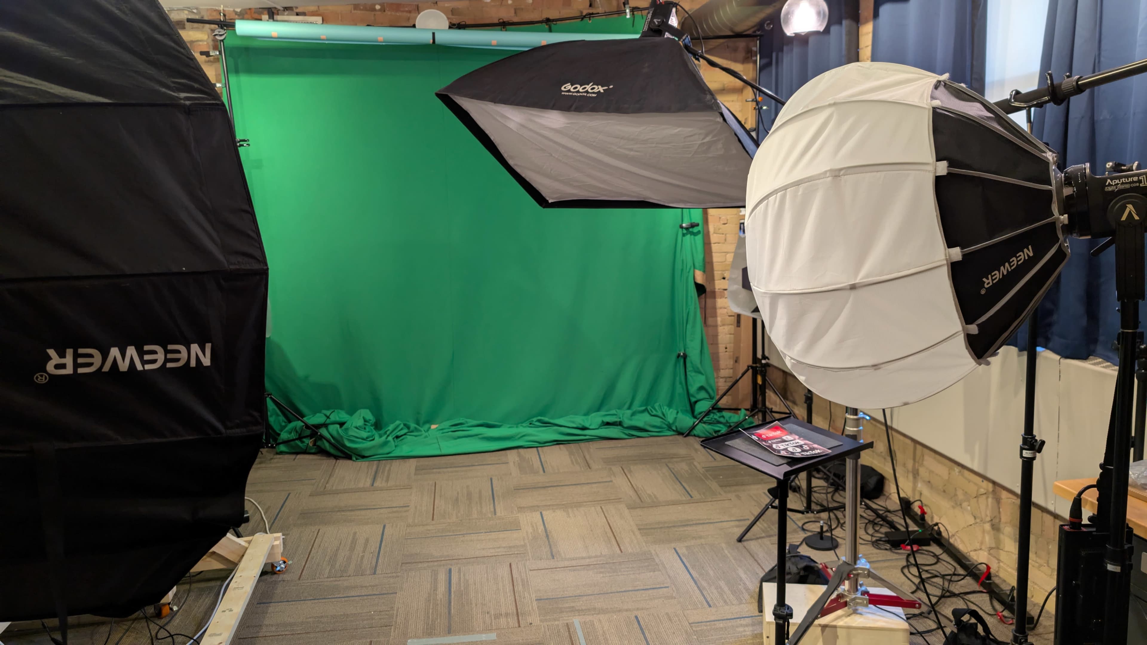 The image shows a photography studio set up with a green backdrop and three lighting setups, including a softbox and an umbrella light, on a tiled floor.