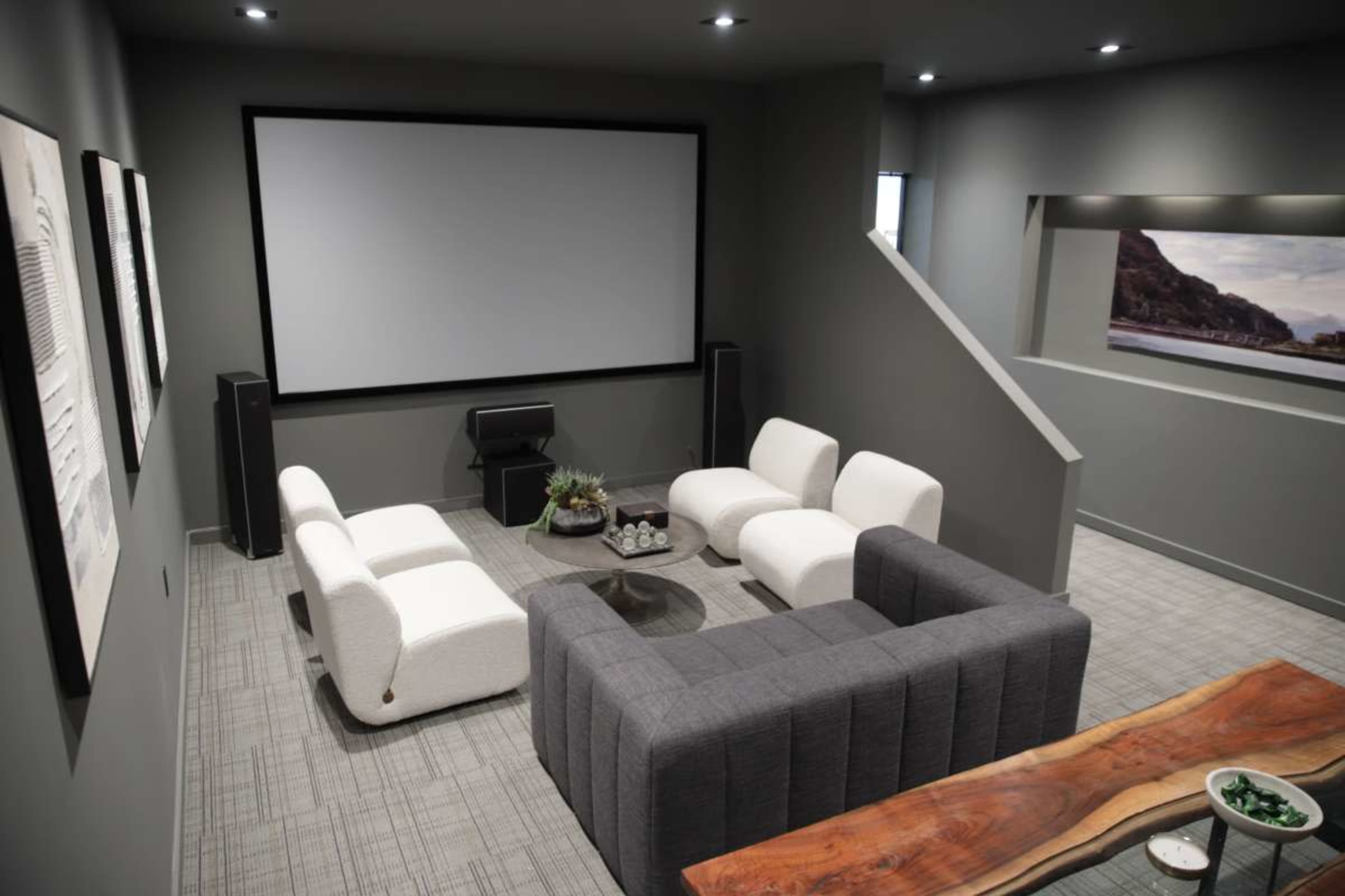 The image shows a modern home theater setup with white and gray seating arranged around a central coffee table, facing a large projection screen.