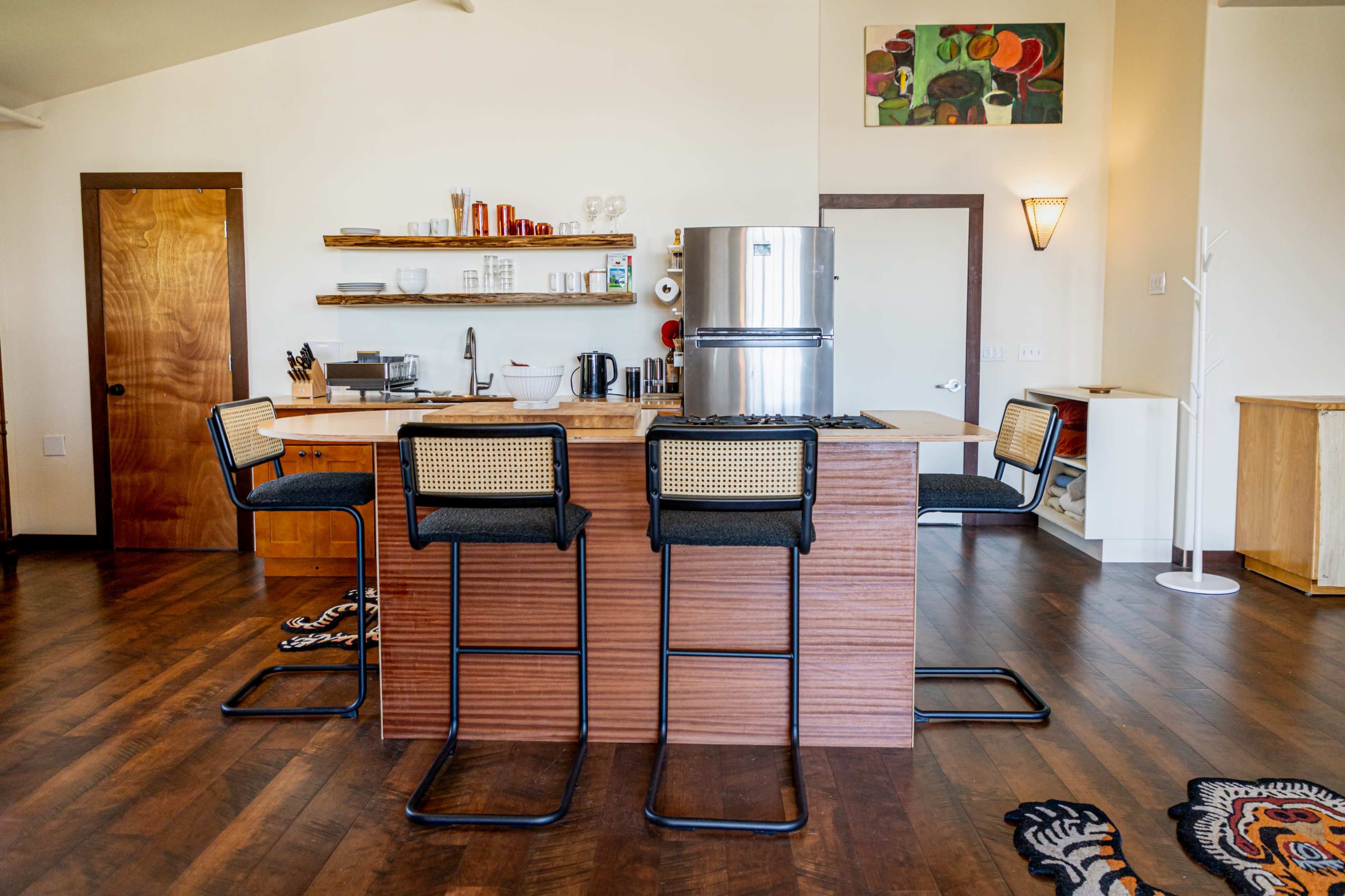 The image shows a modern kitchen with a wooden island, four black bar stools, and stainless steel appliances, featuring shelves of kitchenware and artwork on the wall.
