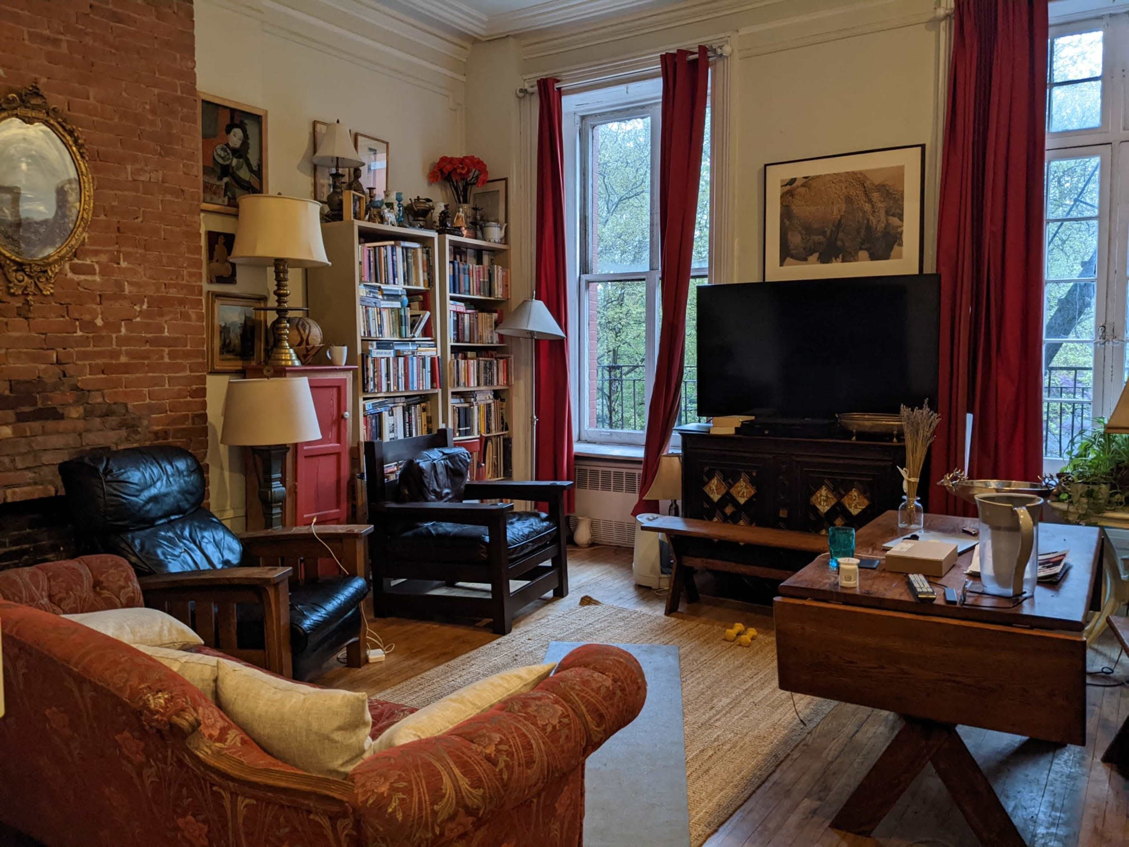 The living room features a mix of seating, a large bookshelf, a television, and a window with red curtains, set against a backdrop of exposed brick walls.