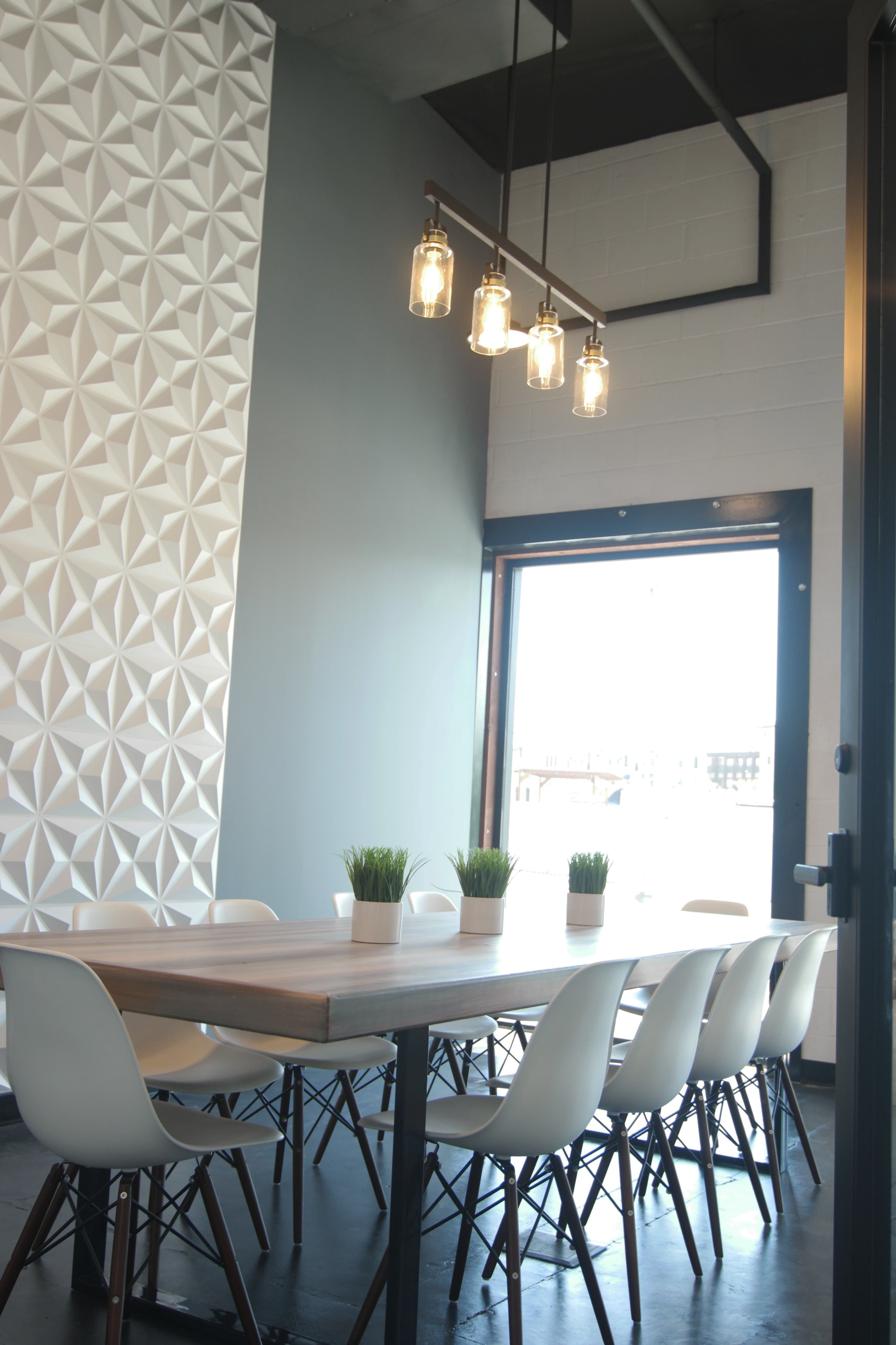 A modern meeting room features a wooden table surrounded by white chairs, with potted plants and pendant lighting above.