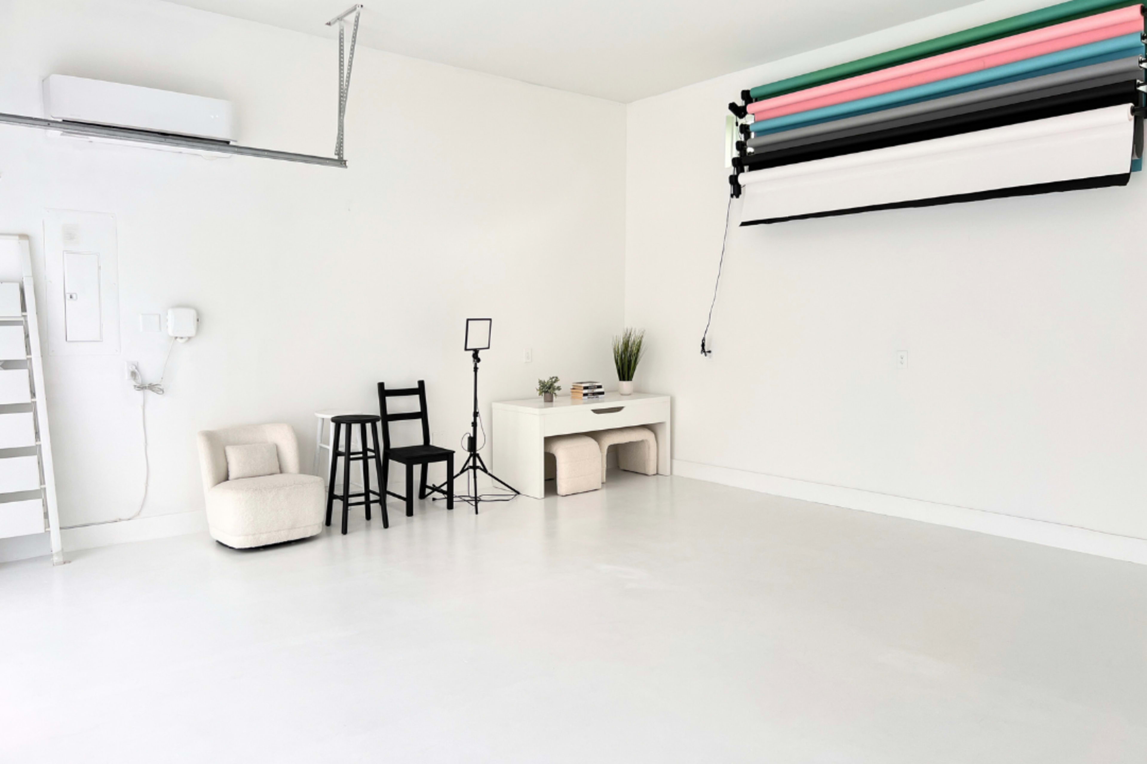 The image shows a minimalist studio space with white walls and a smooth floor, featuring a chair, a small table with a plant, a light stand, and a roll of backdrops mounted on the wall.