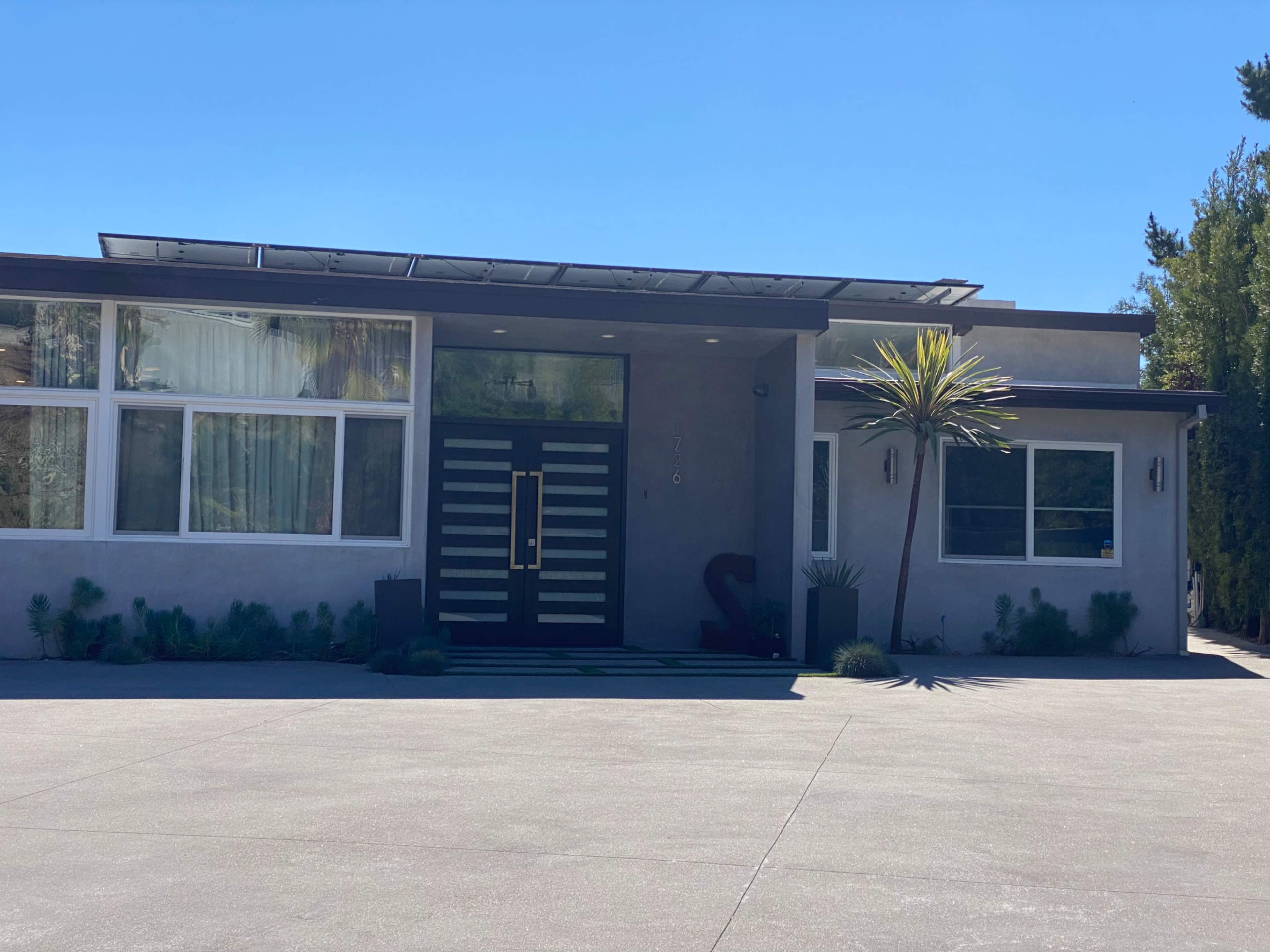 A modern house entrance with a large front door featuring horizontal stripes, flanked by a palm plant and a driveway.