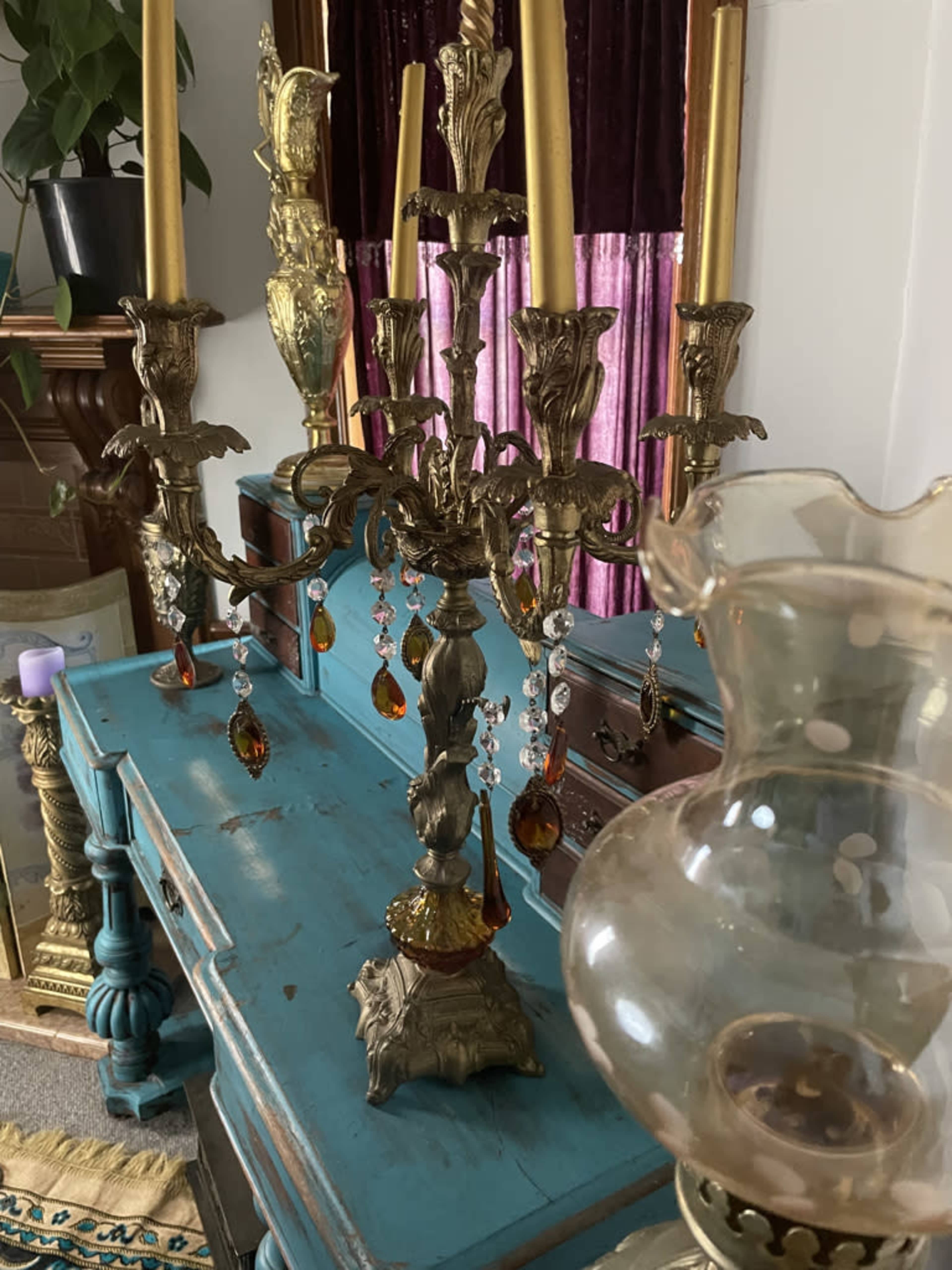 A decorative brass candelabrum with hanging crystals and four tall candle holders stands on a turquoise table, alongside an antique oil lamp.