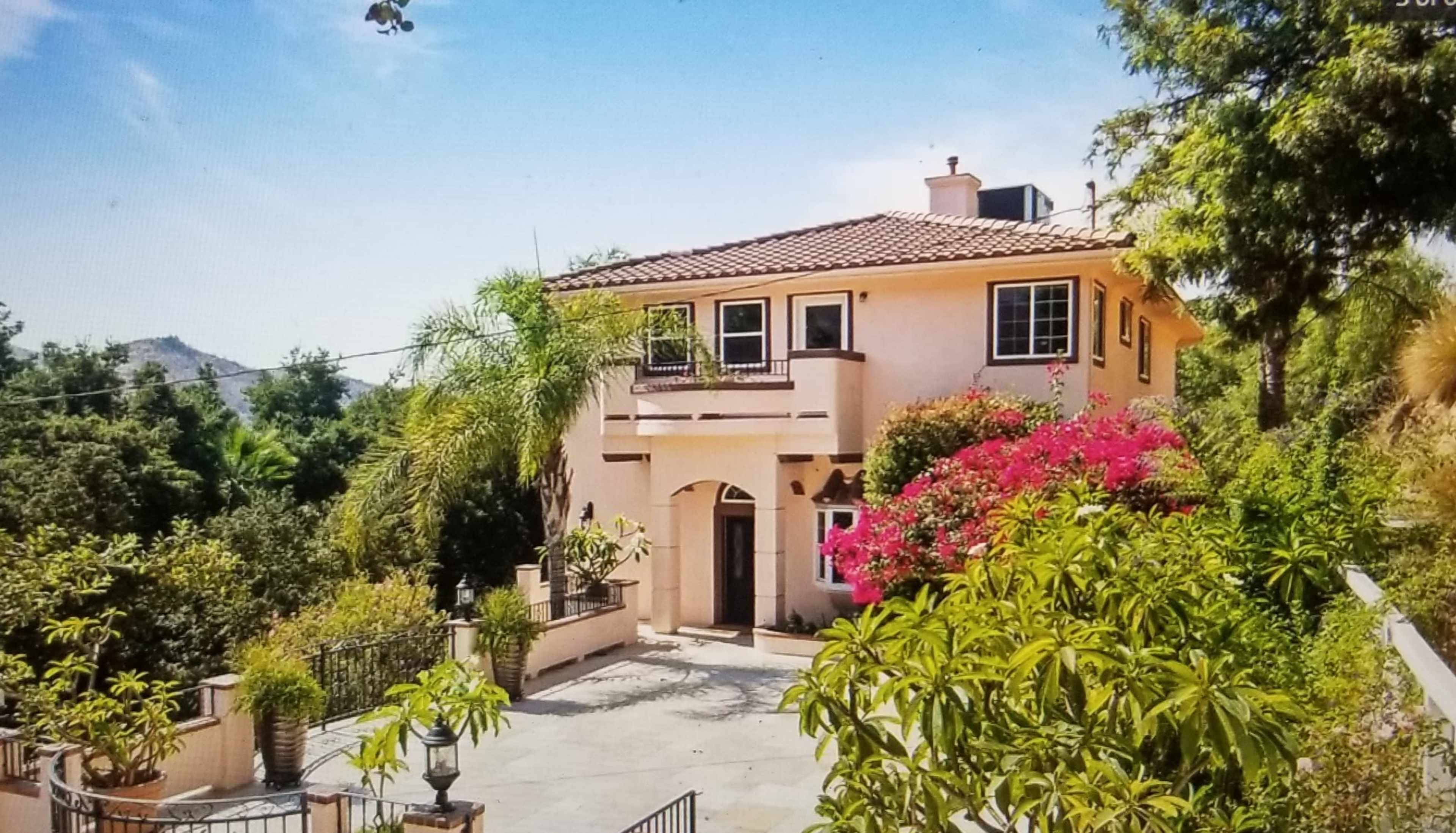 A two-story house with a terracotta roof is surrounded by lush greenery and colorful flowers.